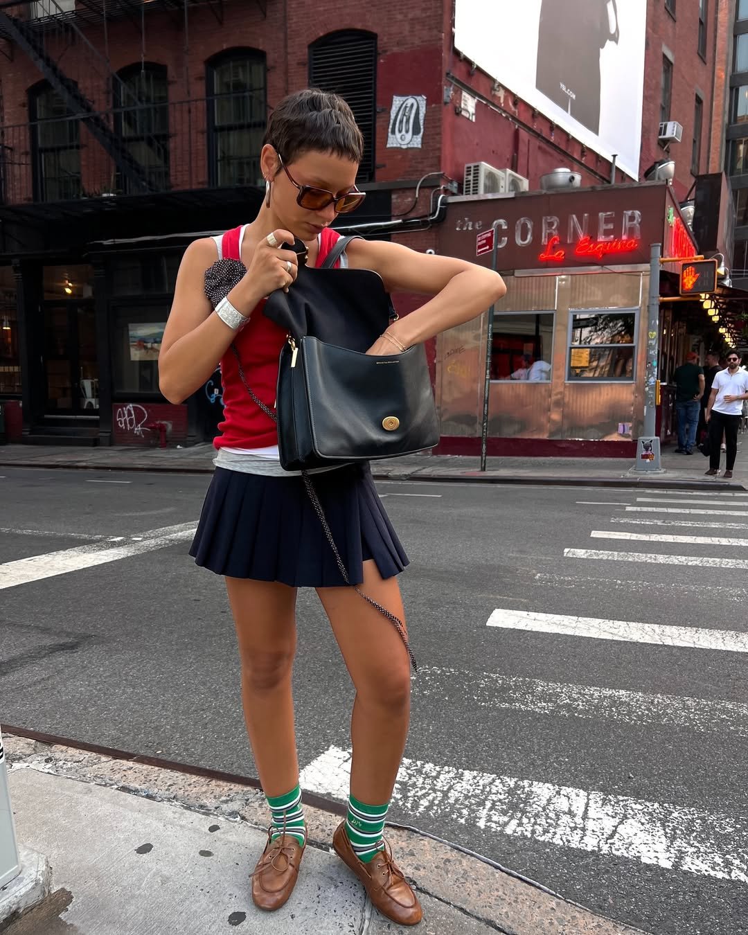 Pessoa em estilo preppy, usando saia plissada, blusa vermelha e óculos de sol, busca algo na bolsa em esquina urbana.