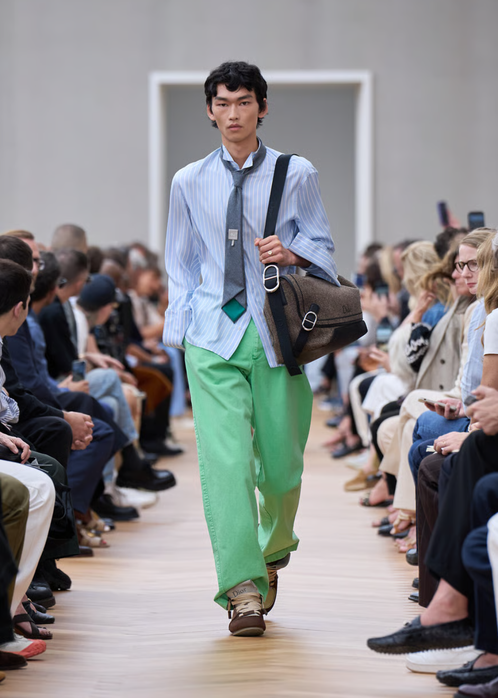 Modelo desfilando com look preppy: terno azul listrado, gravata cinza, calças verdes e bolsa marrom, cercado por espectadores atentos.