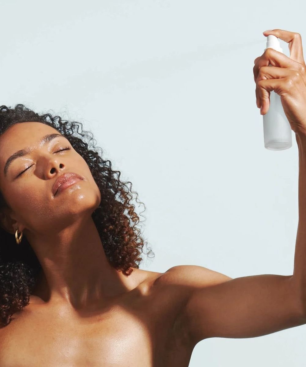 Mulher com olhos fechados borrifa água micelar no rosto, transmitindo sensação de frescor e cuidado com a pele.