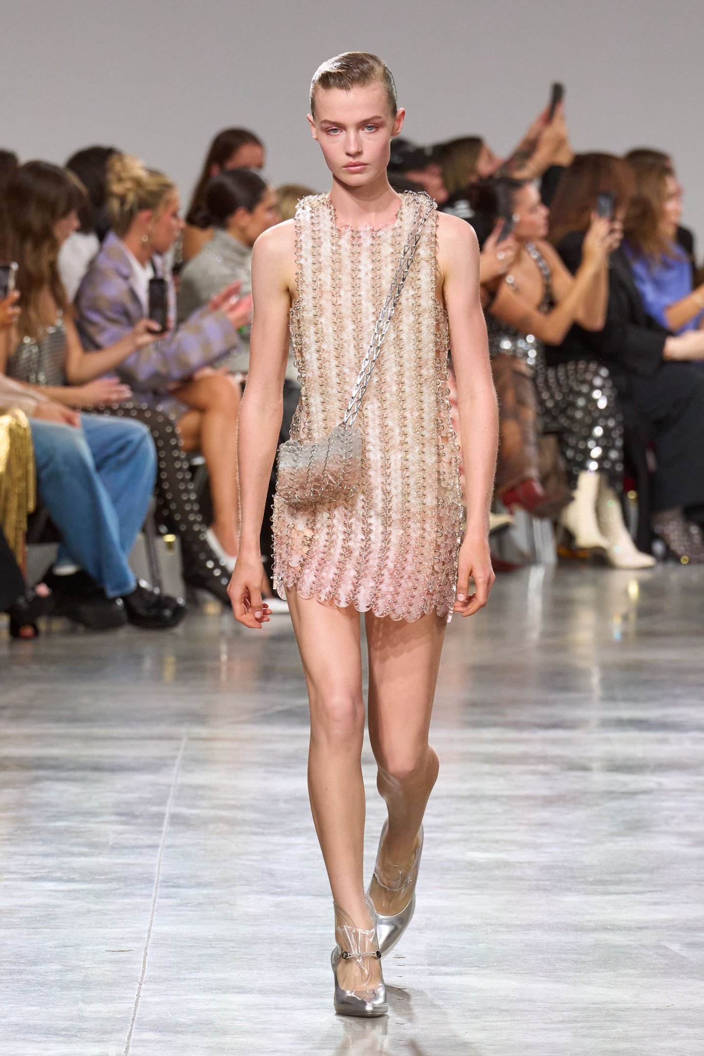 Desfile de moda com modelo usando vestido brilhante na passarela, público ao fundo. Sem referência direta à moda dos piercings.