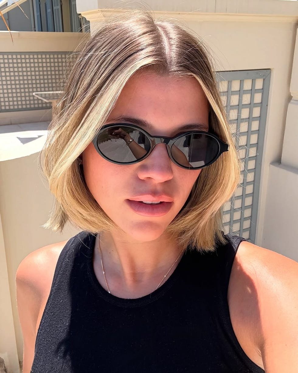 Pessoa com cabelo curto e óculos escuros, estilo riviera bob, vestindo regata preta em ambiente iluminado.