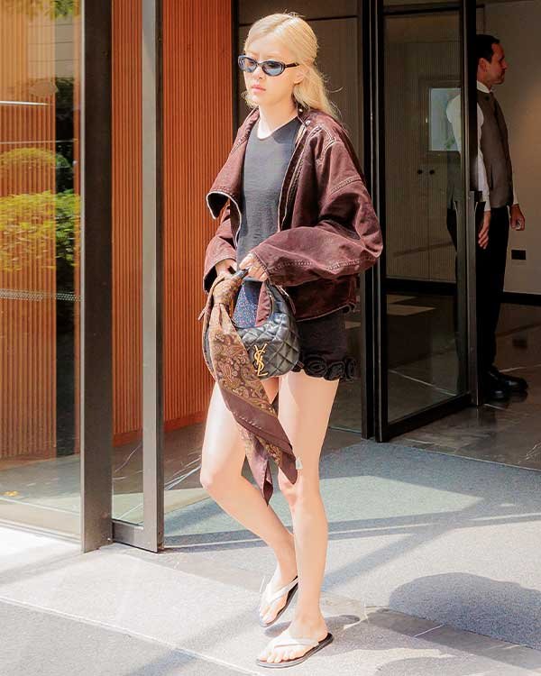 Pessoa saindo de um prédio, usando jaqueta marrom e óculos escuros, carregando uma bolsa Icarino da Saint Laurent.