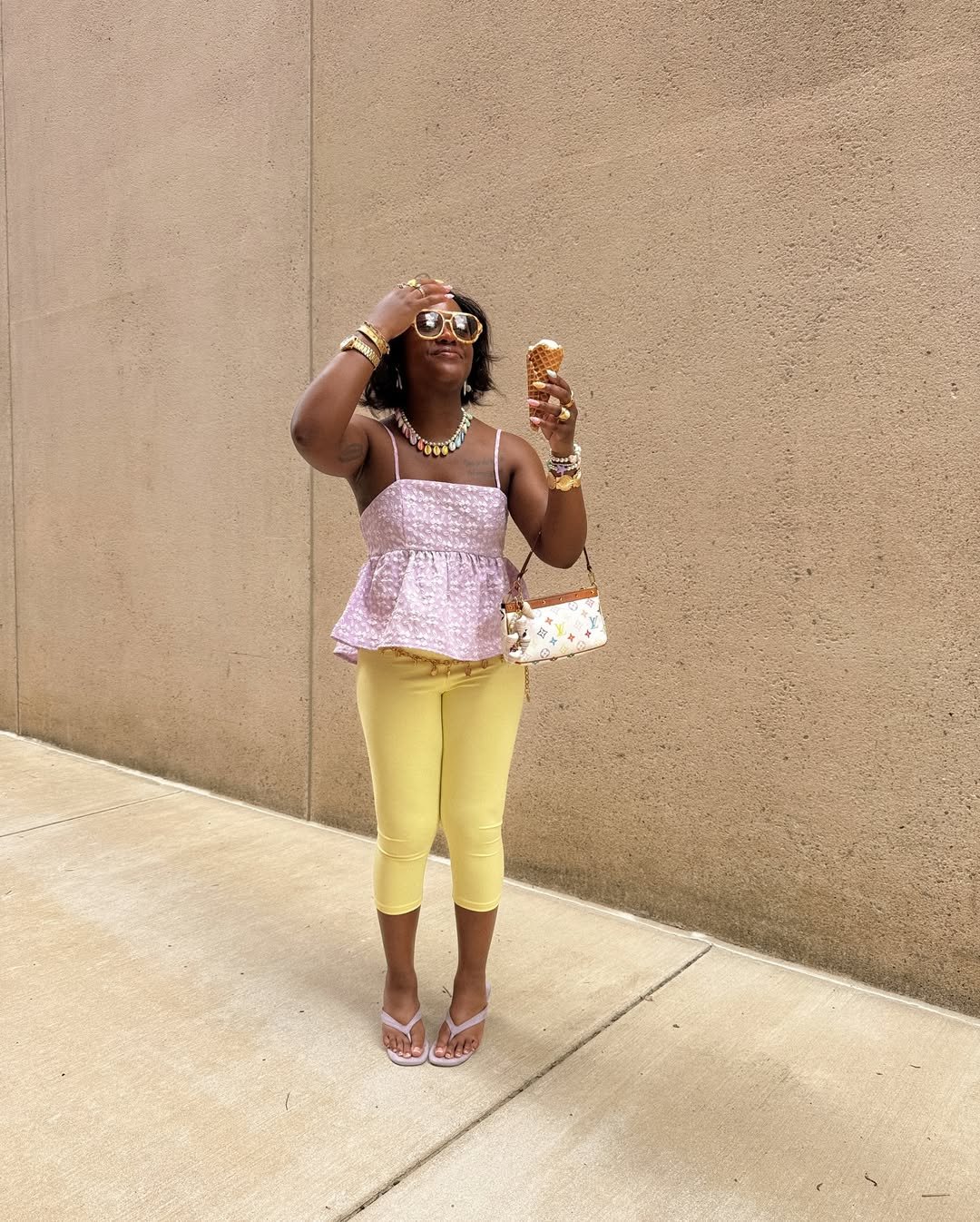 Pessoa com roupas coloridas, top lilás, calça amarela e acessórios, segurando sorvete em frente a parede bege.