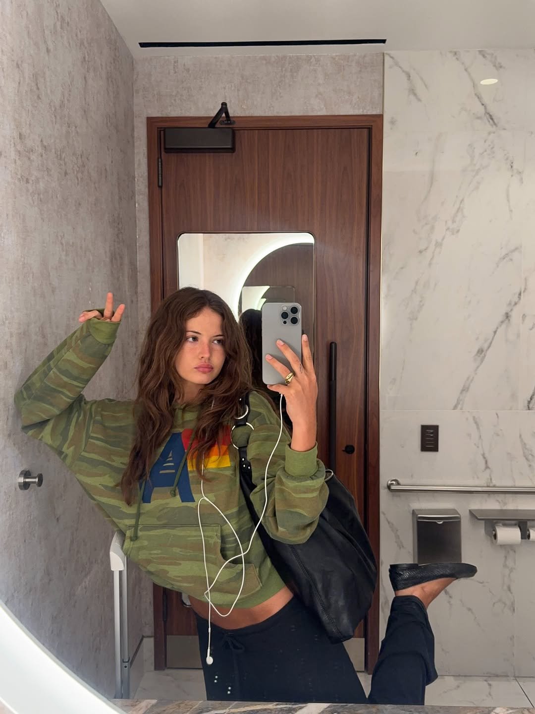 Usuário com moletom camuflado tira selfie no banheiro, posando descontraidamente.