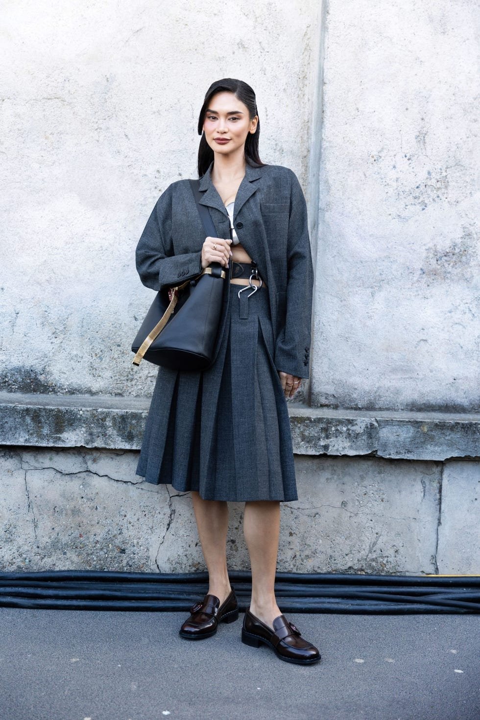 Pessoa vestindo saia tendência de pregas cinza com blazer combinando, bolsa preta e mocassins.