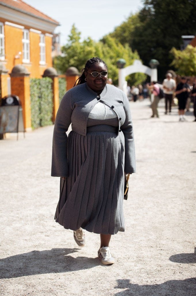 Pessoa usando saia cinza de pregas e jaqueta combinando, andando em local ensolarado.