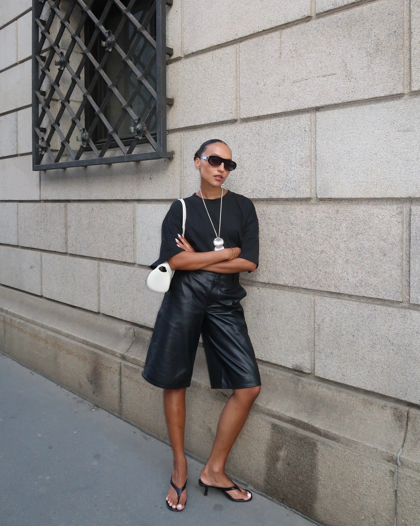 Pessoa posando na rua com camiseta preta, bermuda de couro e sandália de dedo tendência. Ela carrega uma bolsa pequena.