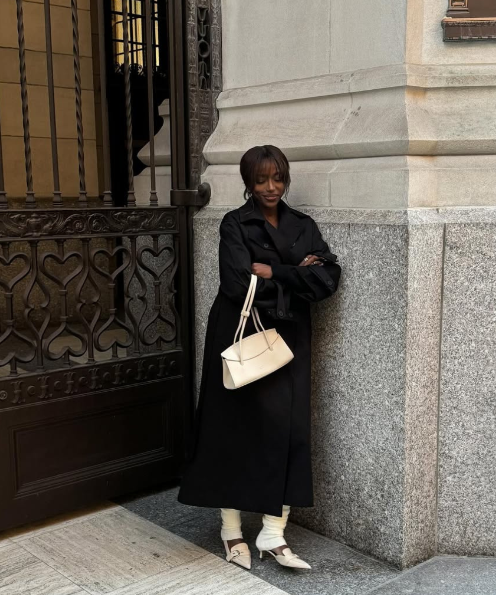 Mulher de casaco preto e sandália feminina branca segura bolsa clara, encostada em portão de ferro decorado.
