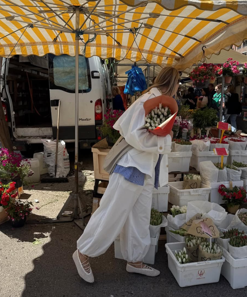 Pessoa em feira ao ar livre, com buquê de flores, vestindo sandália feminina branca e roupa larga.
