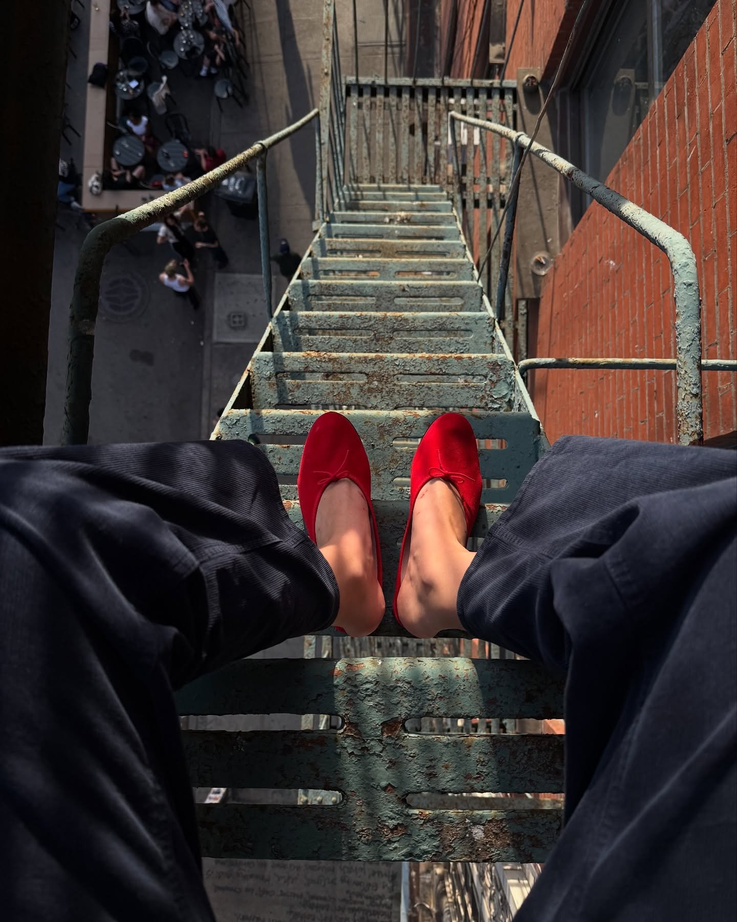 Pessoa sentada em escada de emergência, usando sapatos vermelhos. Abaixo, há uma calçada movimentada.