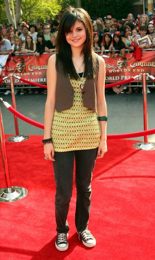 Jovem sorrindo em tapete vermelho, vestindo regata estampada e colete marrom, rodeada por pessoas ao fundo.