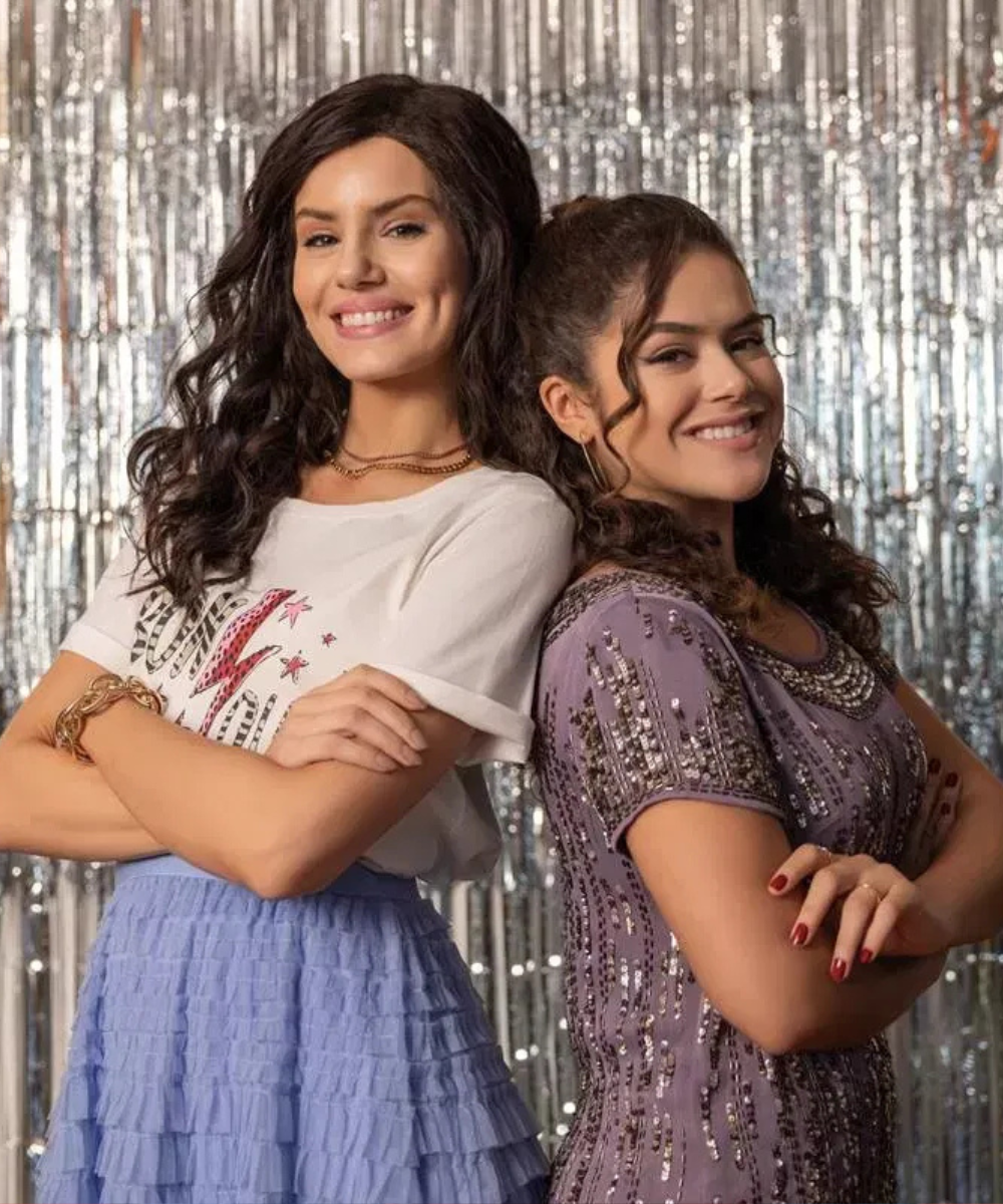 Mulheres sorrindo de braços cruzados, posicionadas lado a lado, em frente a um fundo brilhante. Evento de verão especial.