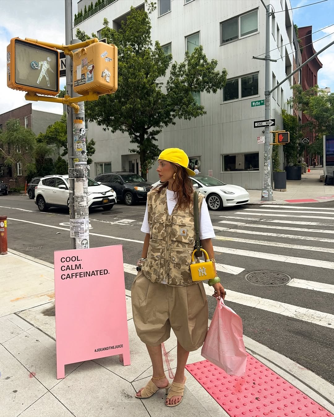 Pessoa com colete camuflado e boné amarelo em cruzamento urbano, segurando bolsa e sacola. Placa rosa ao lado.