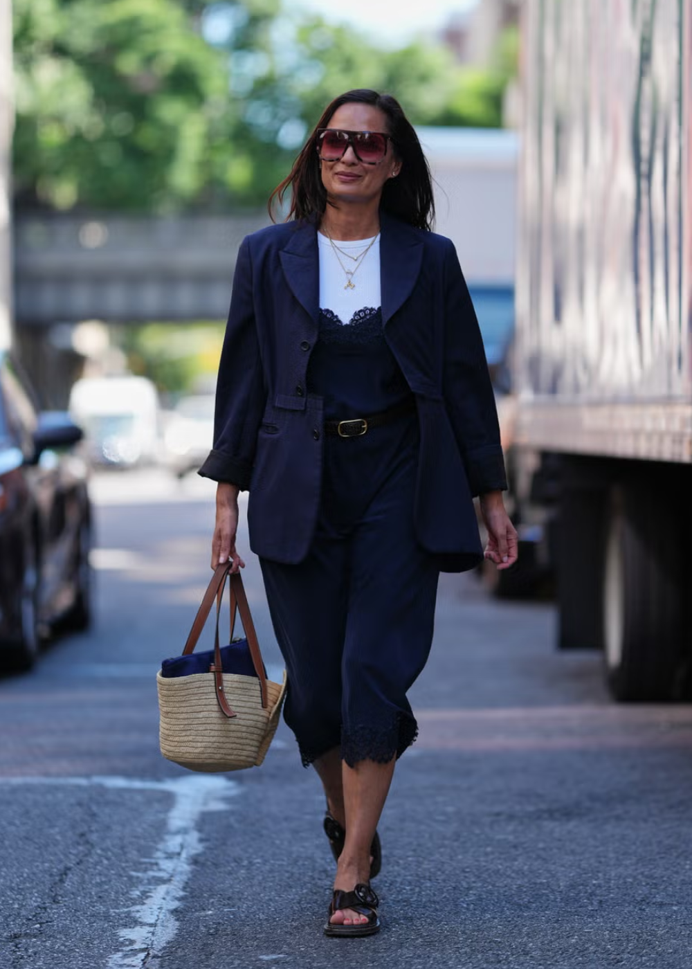 Mulher usando traje azul marinho e óculos escuros, segurando bolsa de palha. Estilo urbano da NYFW.