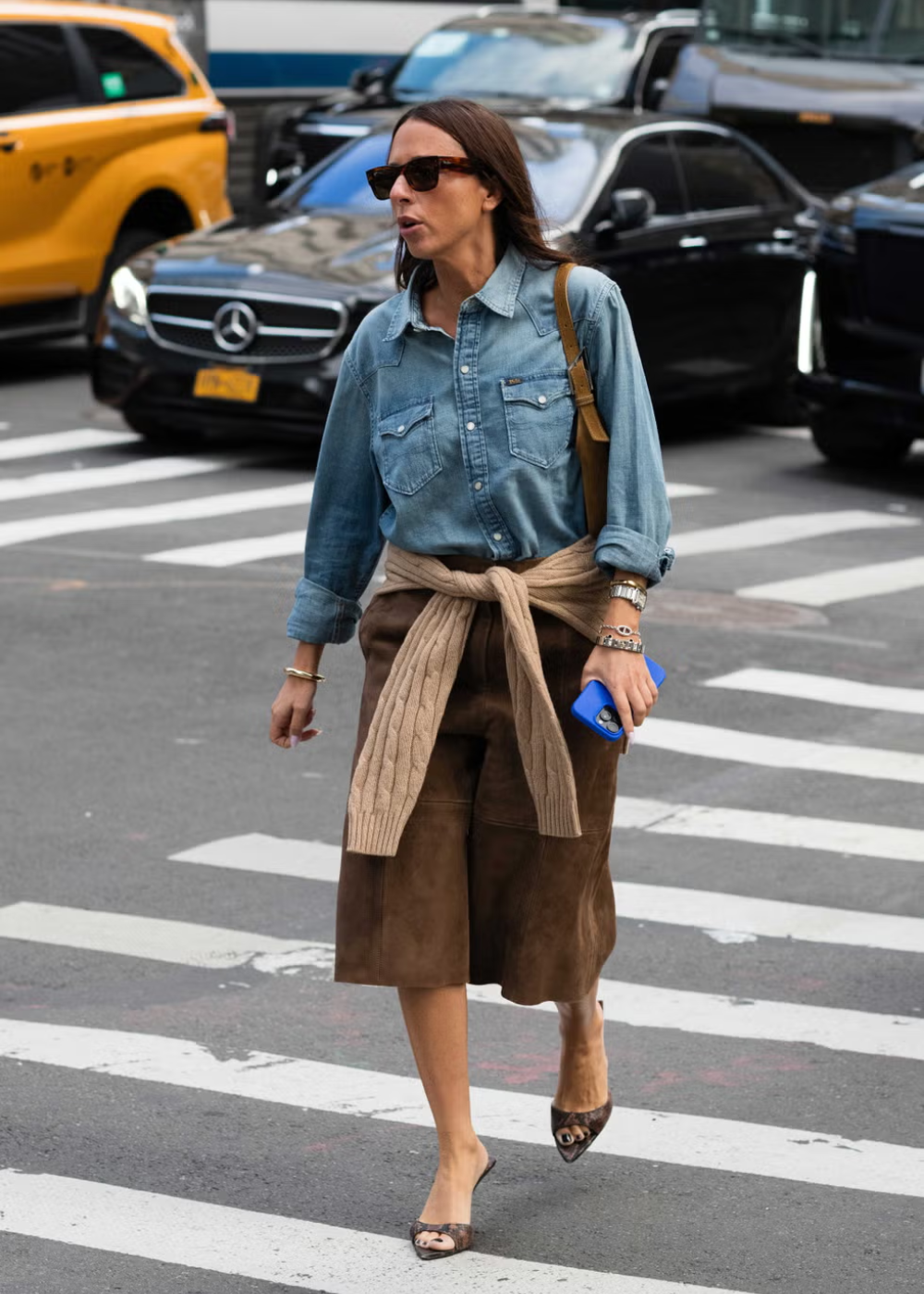 Pessoa com look casual, camisa jeans, e suéter amarrado na cintura. Looks do street style da NYFW, cruzando na faixa.