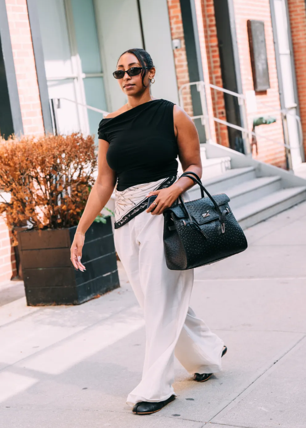 Mulher com óculos escuros usa blusa preta e calça branca larga, carregando bolsa grande; looks do street style da NYFW.