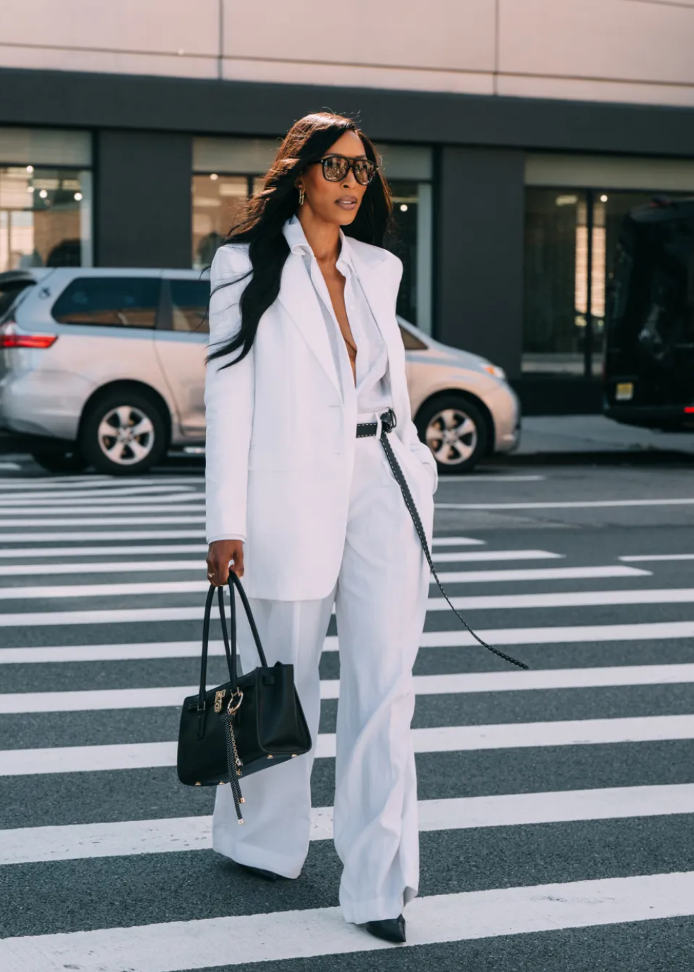 Pessoa atravessa a rua com terno branco elegante e bolsa preta, destacando looks do street style da NYFW.