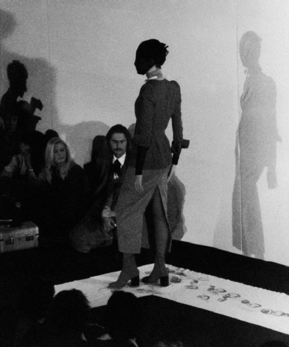 Desfile de moda vintage com pessoa vestindo roupa formal e sombra projetada na parede.
