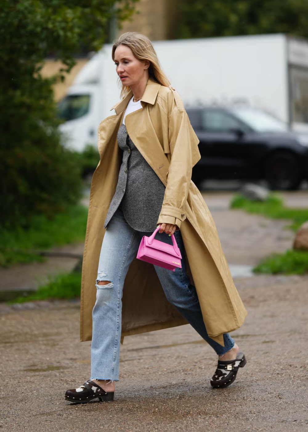 Mulher caminhando com casaco bege, jeans rasgados e bolsa rosa, usando tamancos de madeira escuros.