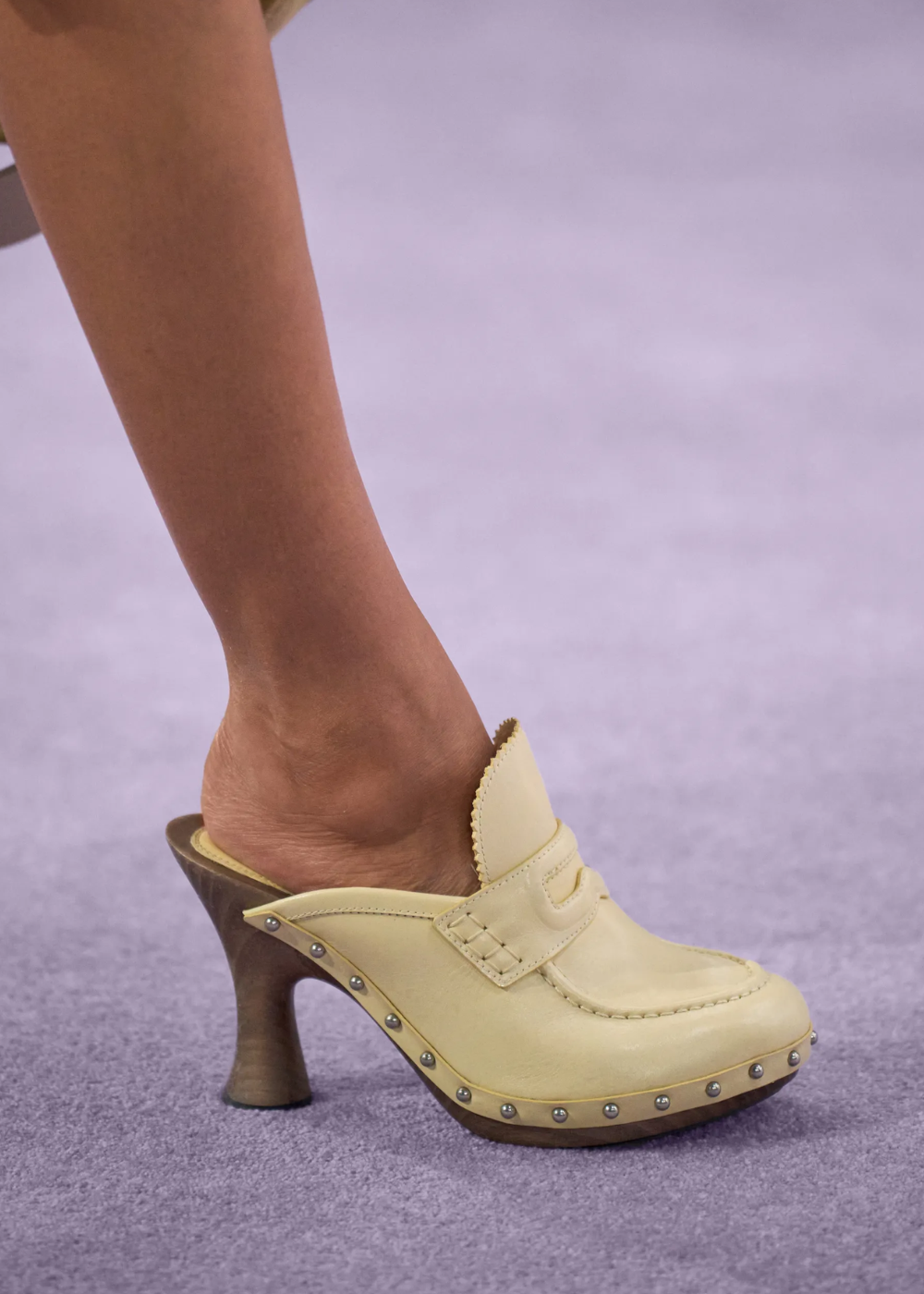 Tamanco de madeira bege com salto alto, decorado com tachas prateadas, em fundo lilás.