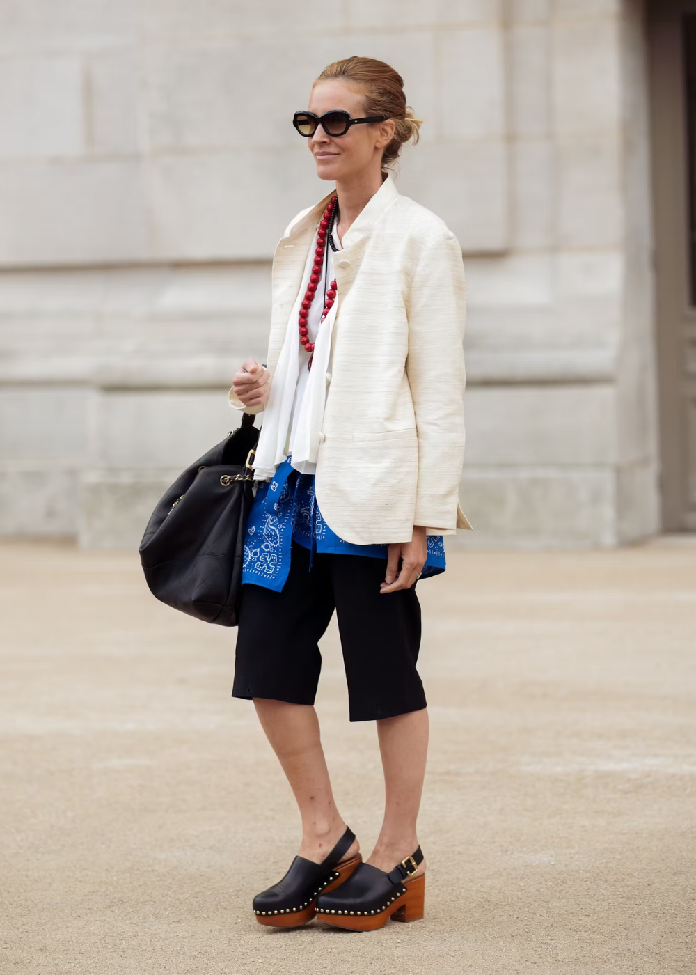 Pessoa usando tamanco de madeira, blusa branca, blazer bege, bermuda preta e colar vermelho, ao ar livre.