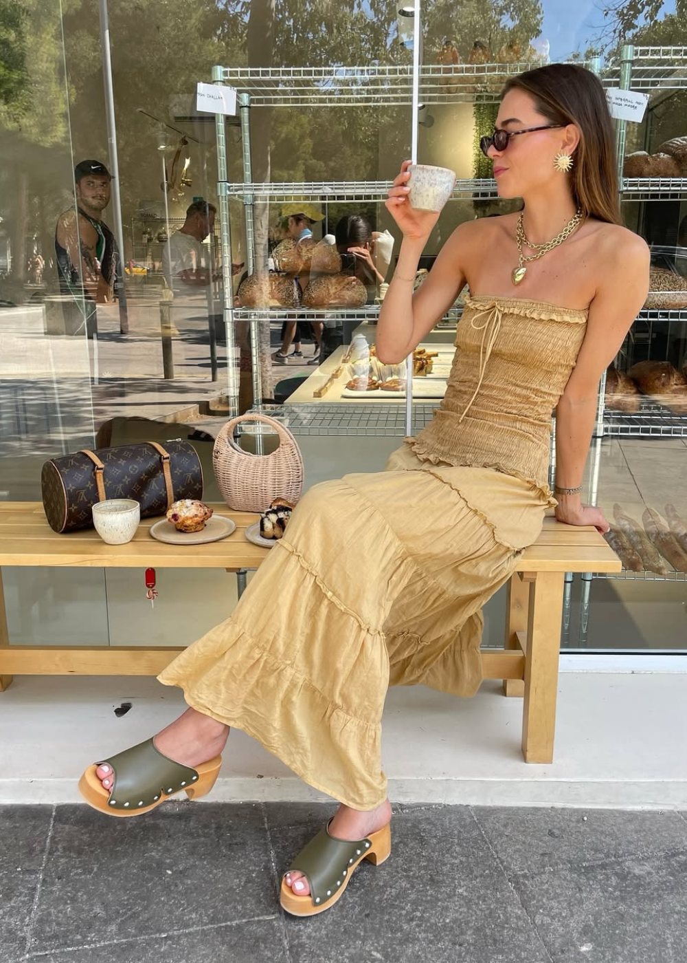 Mulher sentada em banco com vestido bege, tomando bebida. Usa tamancos de madeira e óculos escuros.