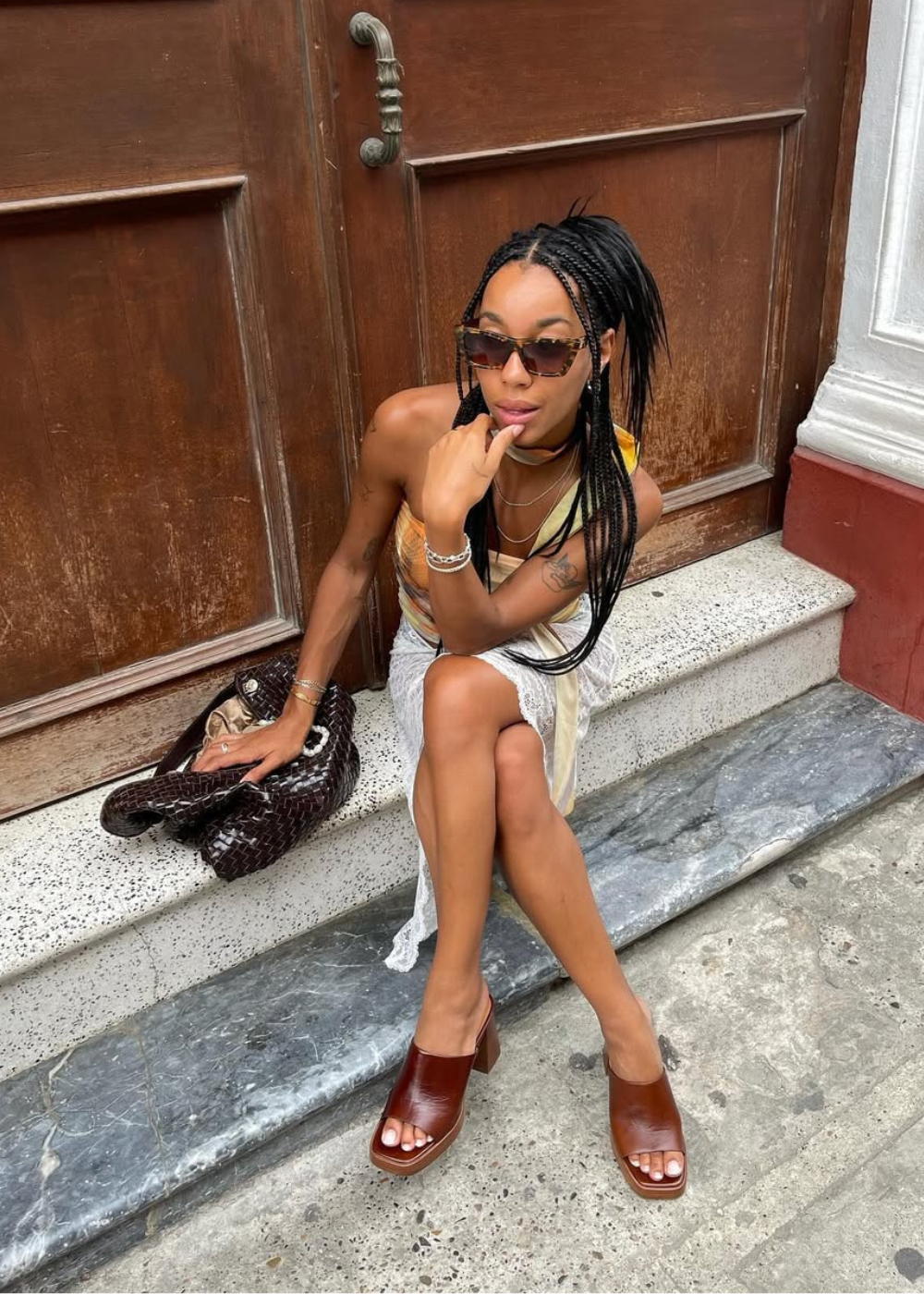 Pessoa sentada em degrau de mármore, vestindo tamanco de madeira marrom, óculos escuros e vestido leve.