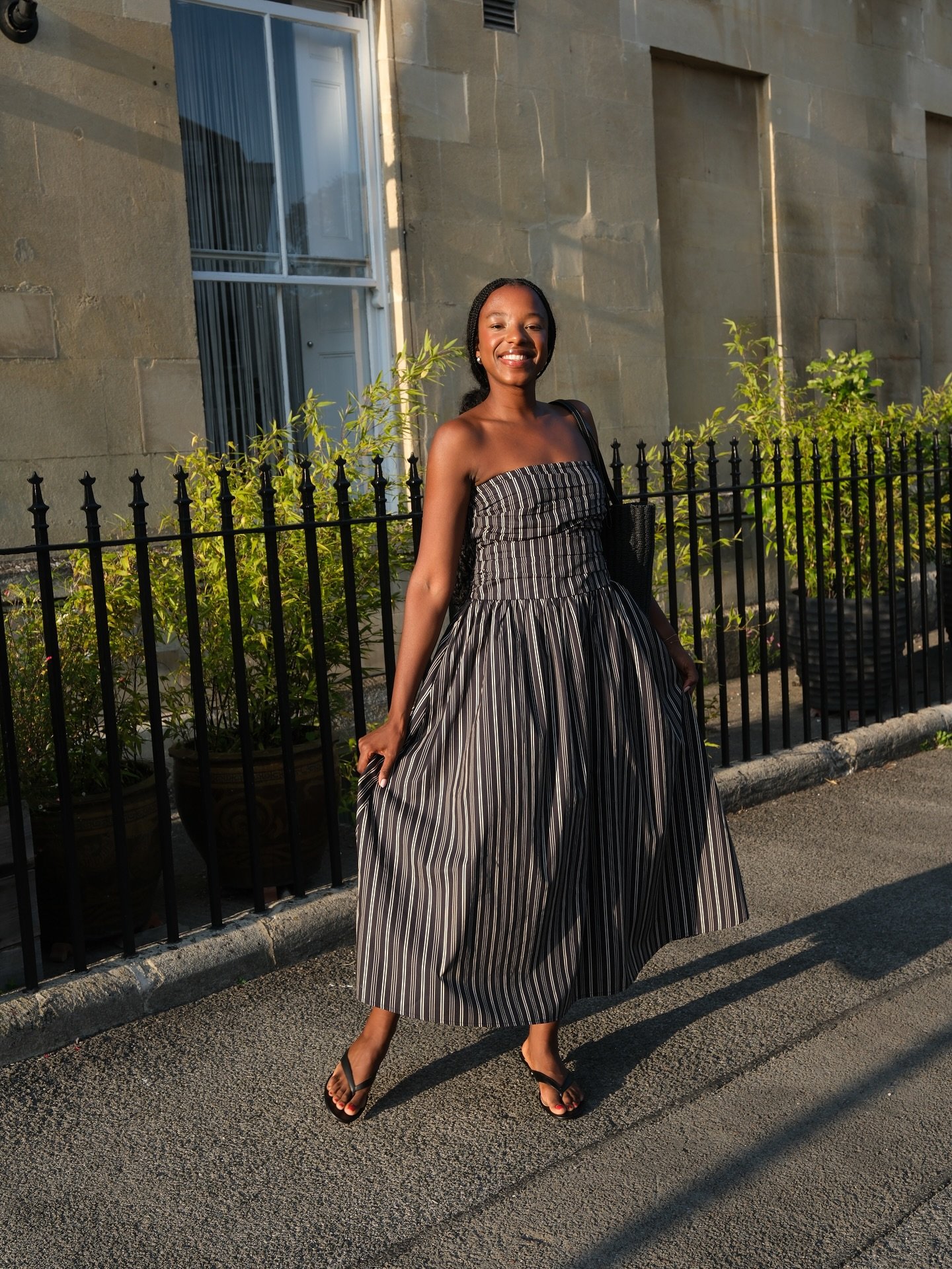 Mulher sorrindo de vestido listrado em ambiente urbano, usando sandália de dedo estilo tendência.