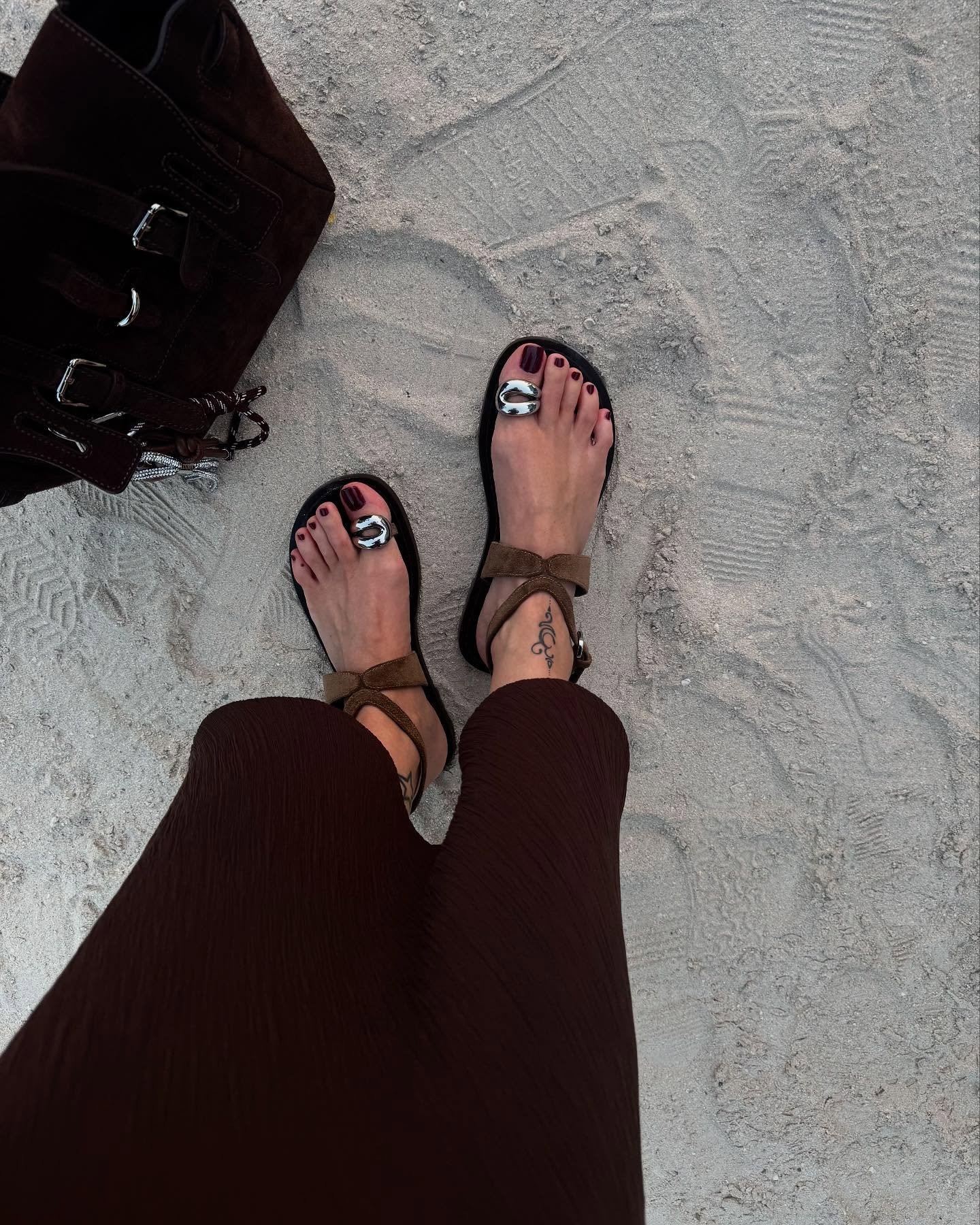 Sandália de dedo com detalhes prateados usada sobre a areia, combinada com bolsa marrom.