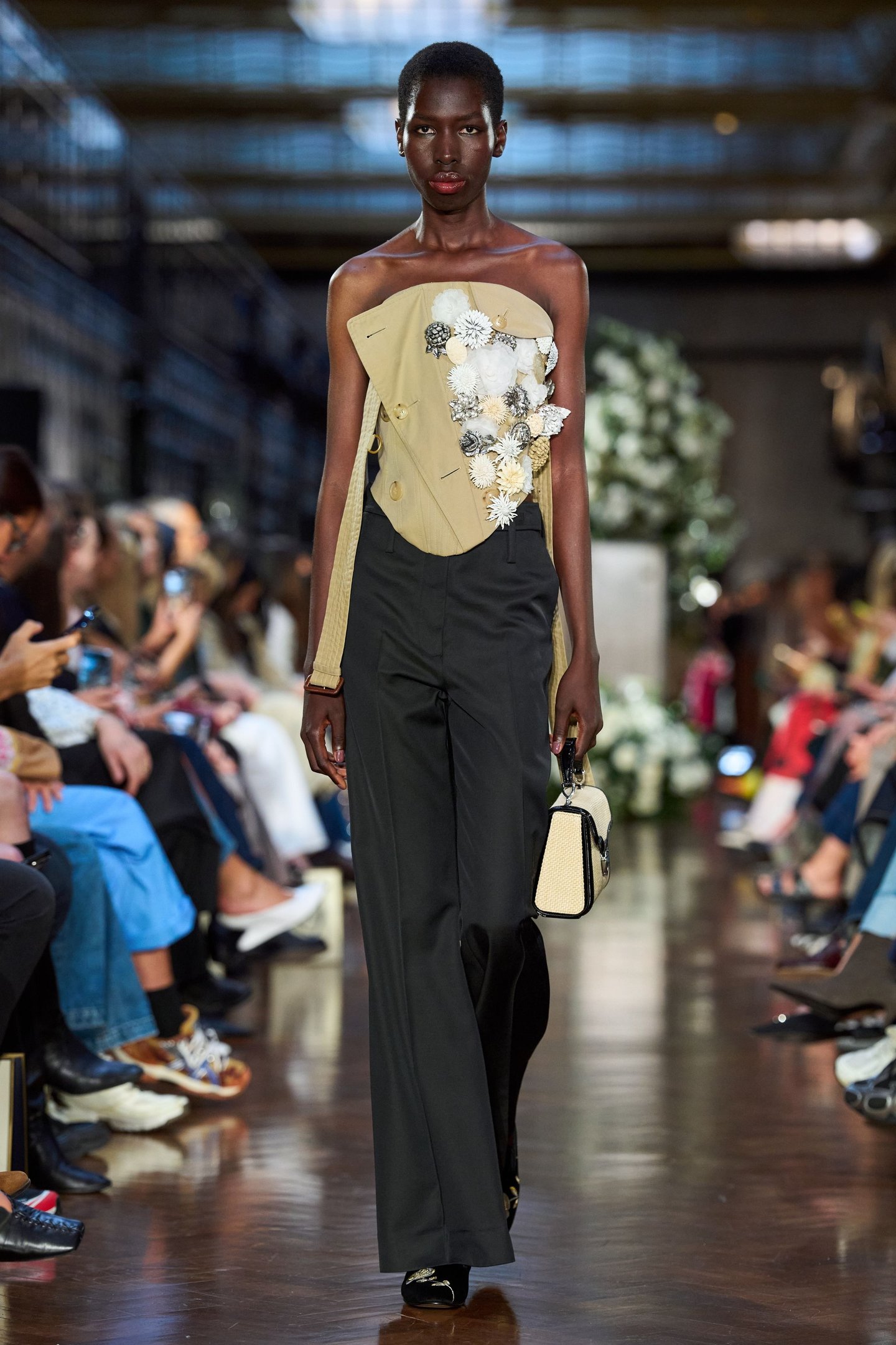 Modelo em desfile, vestindo top bege com flores e calça preta, segurando bolsa na London Fashion Week SS25.