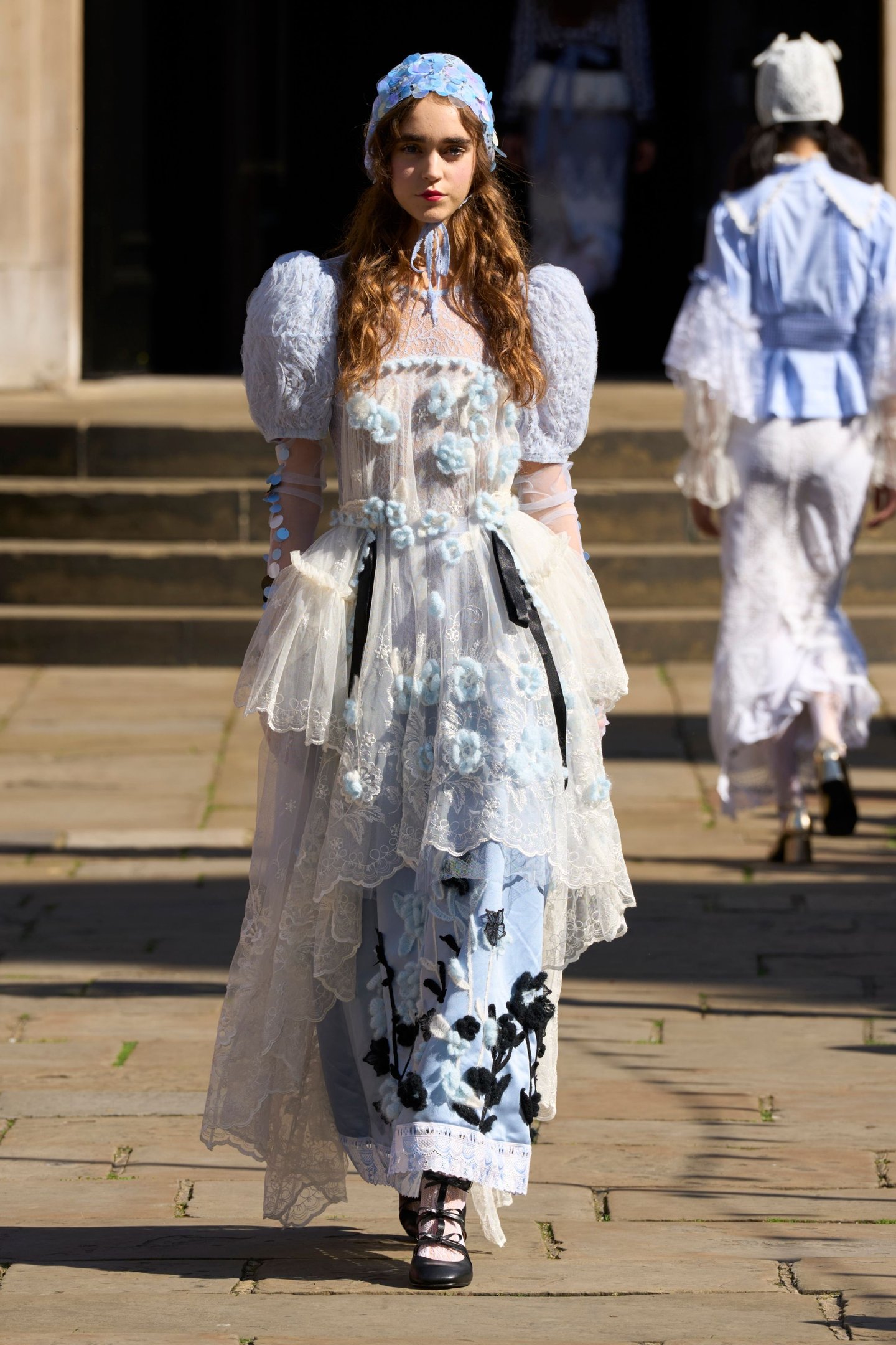 Modelo em passarela vestindo roupa de renda com bordados florais azuis na London Fashion Week SS26.