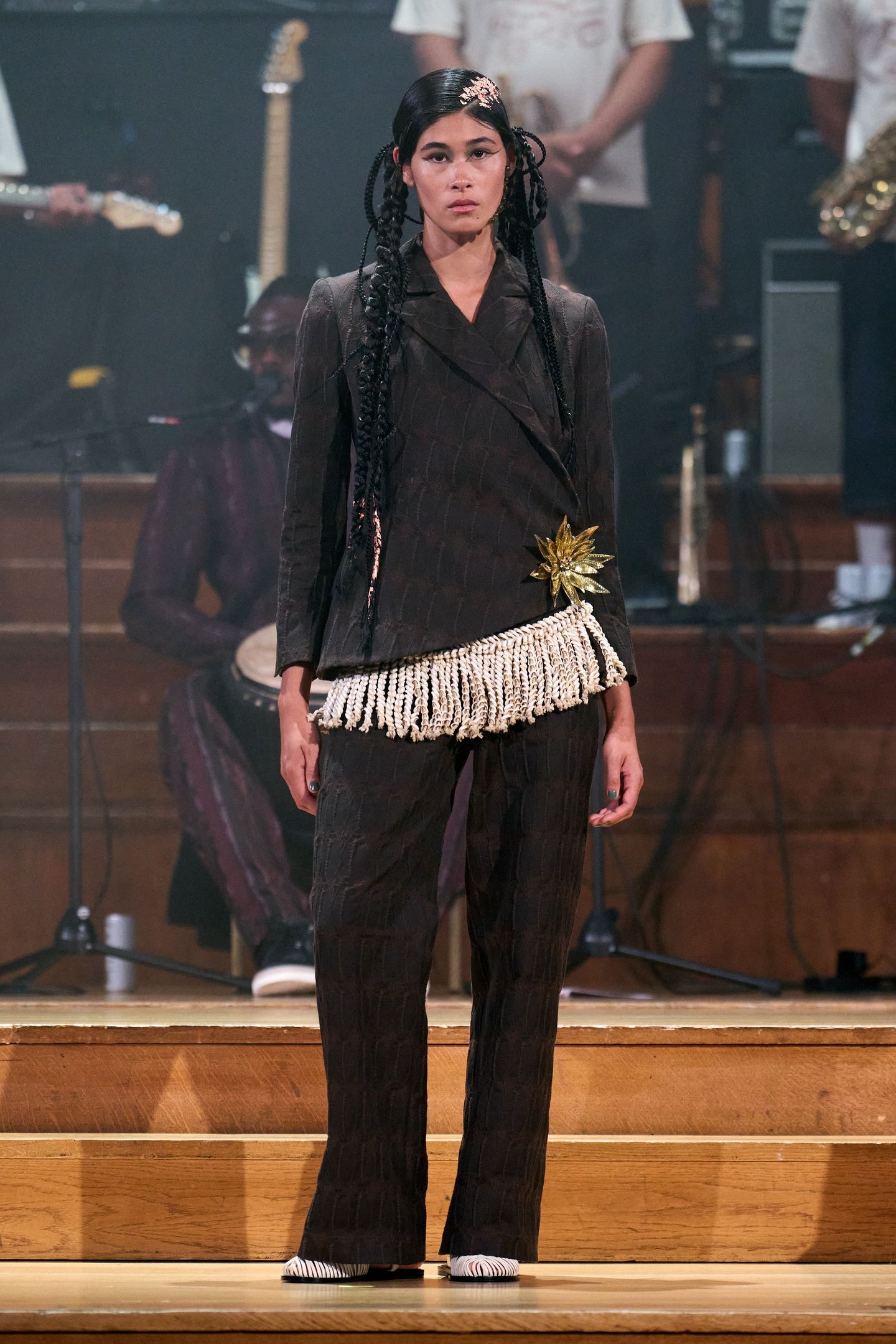 Modelo usando terno escuro com detalhe de franjas e adorno dourado na cintura. Evento inspirado na London Fashion Week SS26.