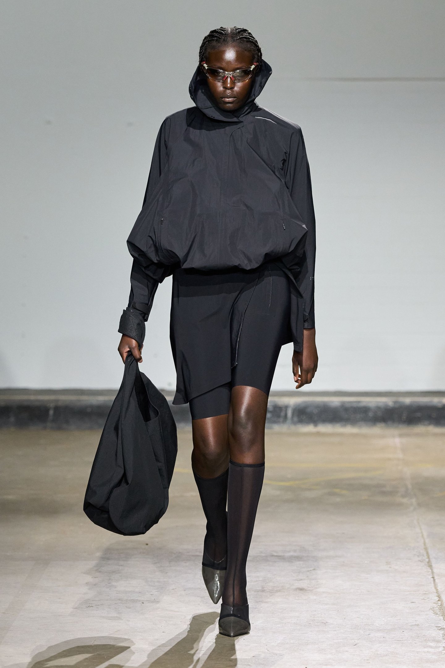 Modelo na passarela com look preto elegante, tênis transparentes e óculos modernos. Evento London Fashion Week SS26.