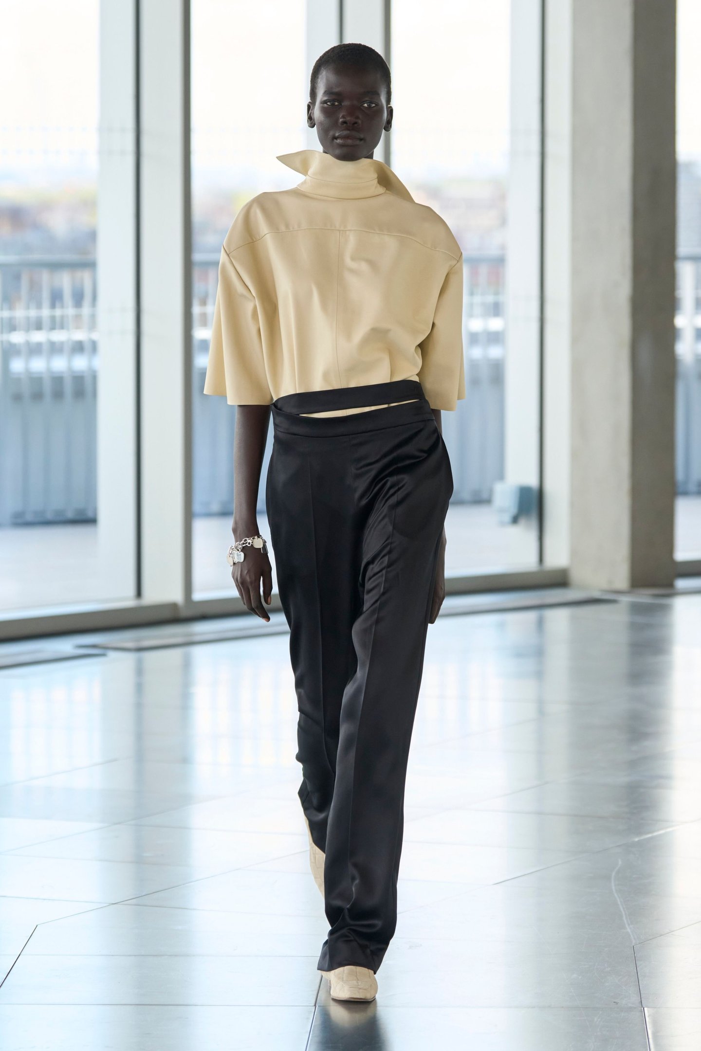 Modelo desfila com blusa bege e calça preta na London Fashion Week SS26, em ambiente moderno.