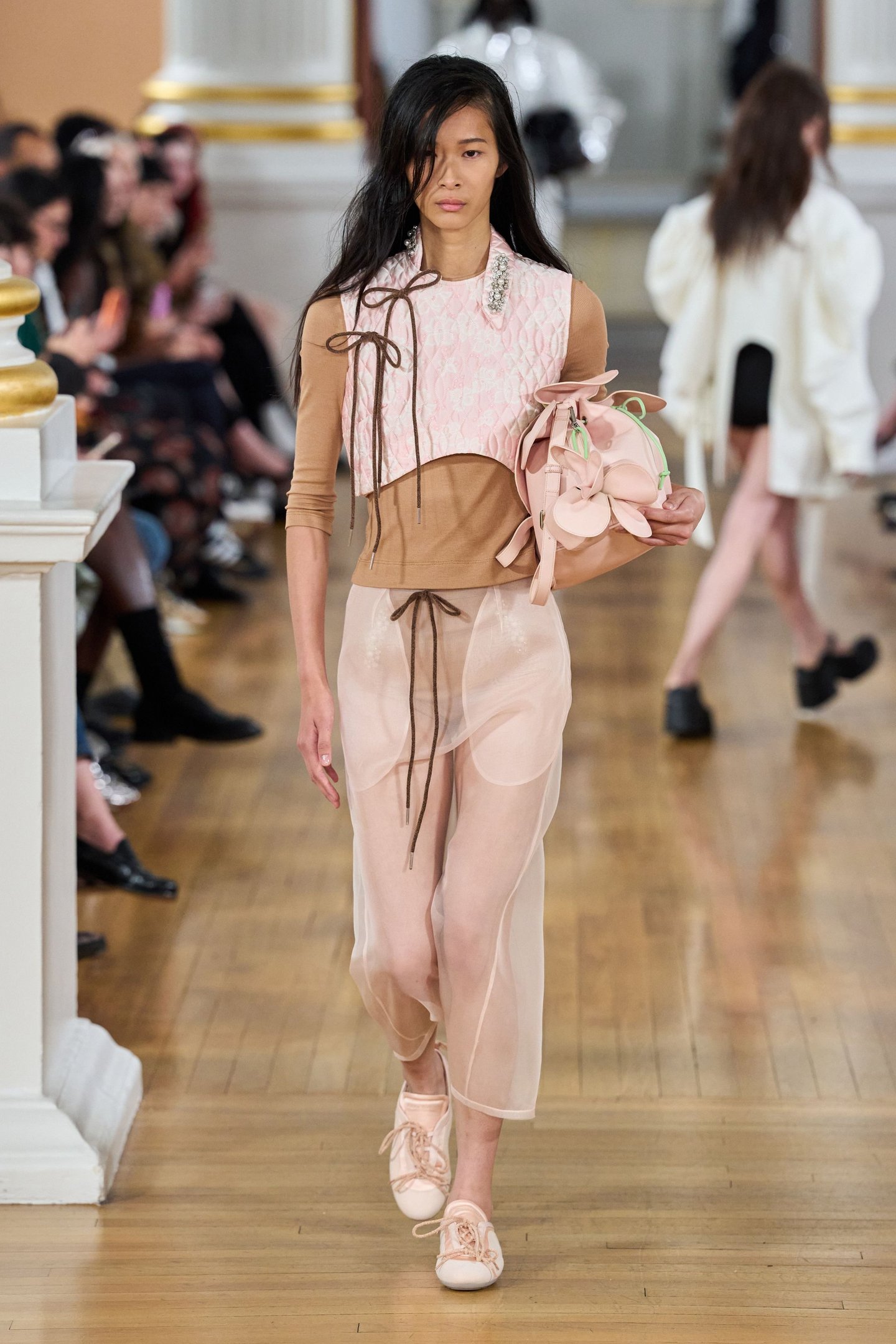 Modelo caminha em passarela usando roupa bege e rosa transparente, carregando bolsa floral em desfile London Fashion Week SS26.