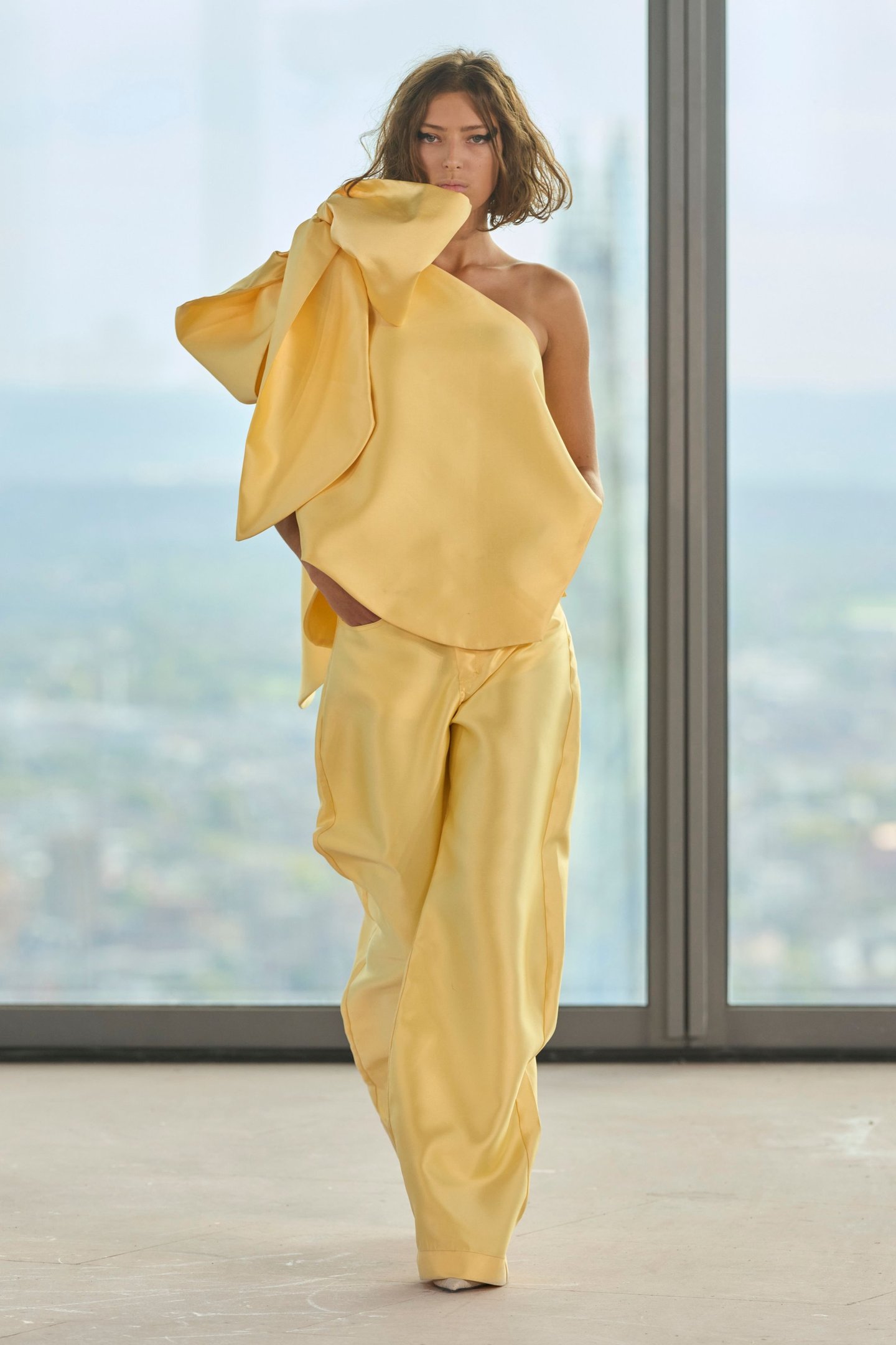Modelo veste conjunto amarelo com top assimétrico e laço na London Fashion Week SS26. Fundo com janela de vidro.