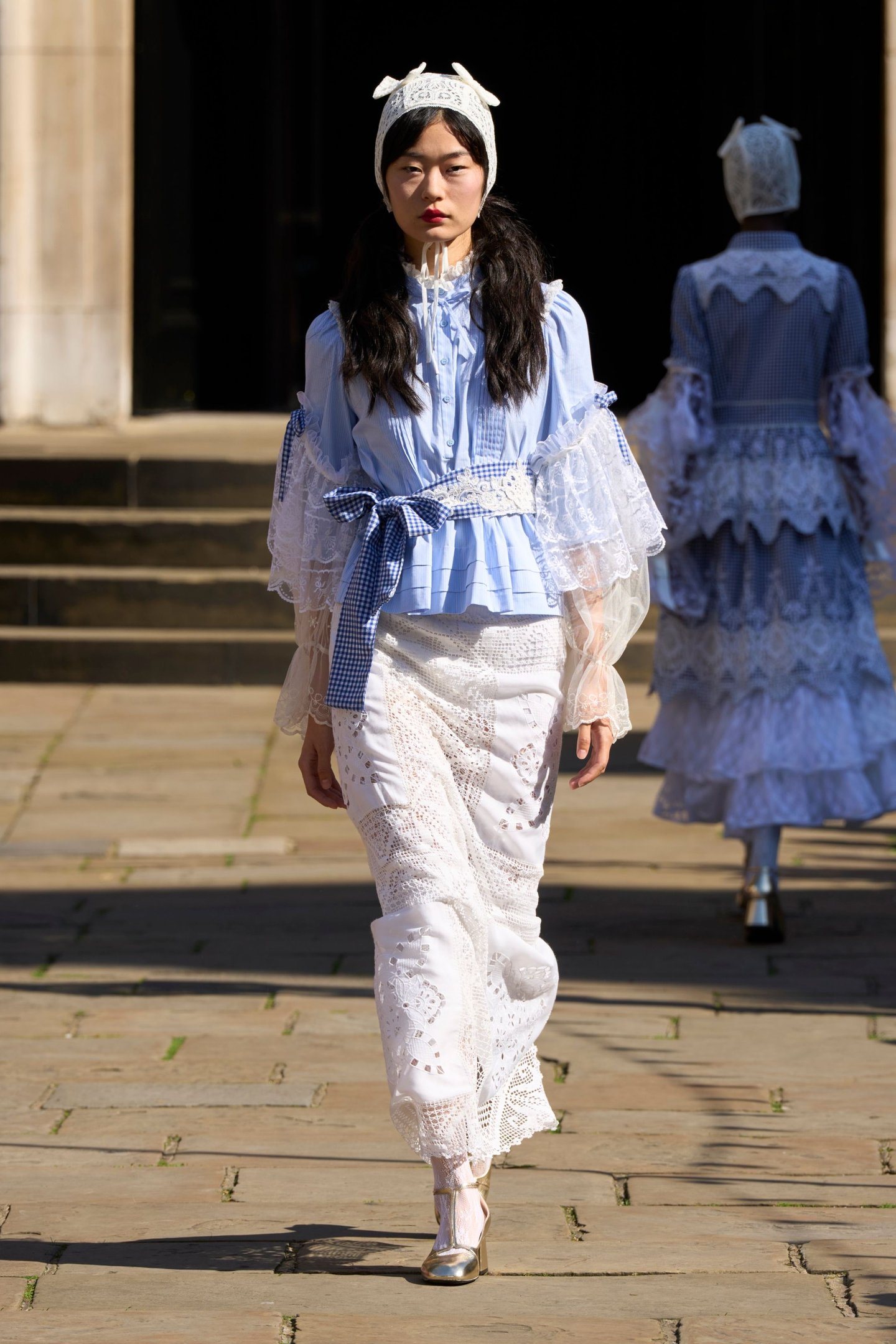 Modelo em passarela com vestido azul e branco rendado, na London Fashion Week SS26. Fundo com escadas.