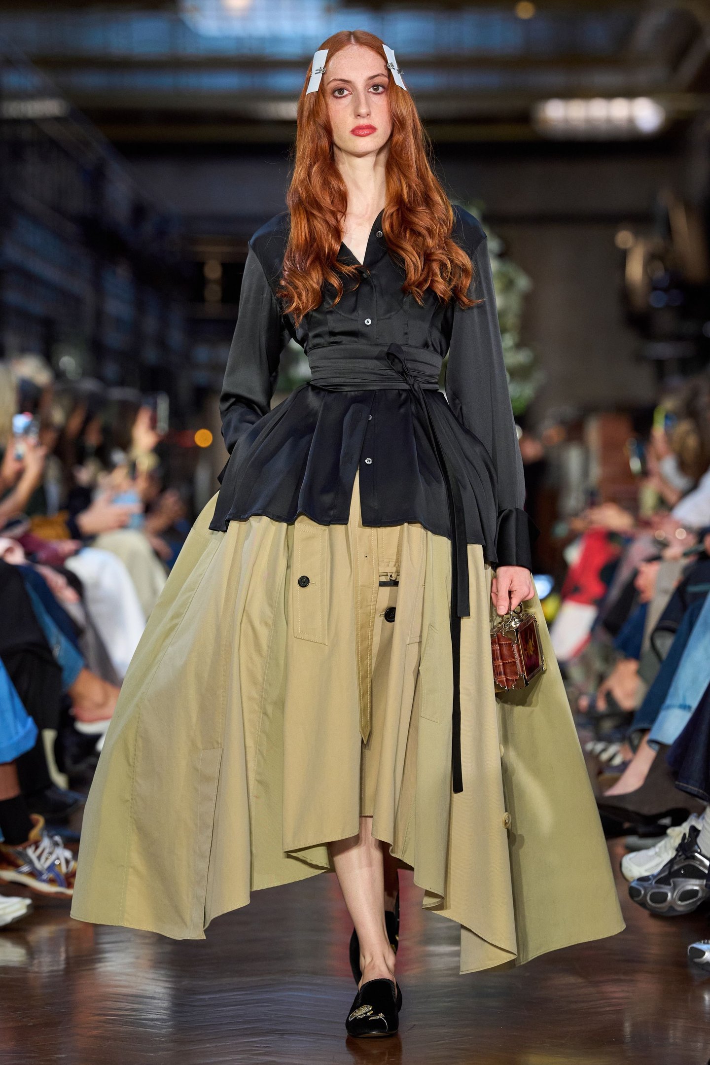 Modelo desfilando na London Fashion Week SS26, usando vestido preto com saia bege assimétrica e segurando bolsa pequena.