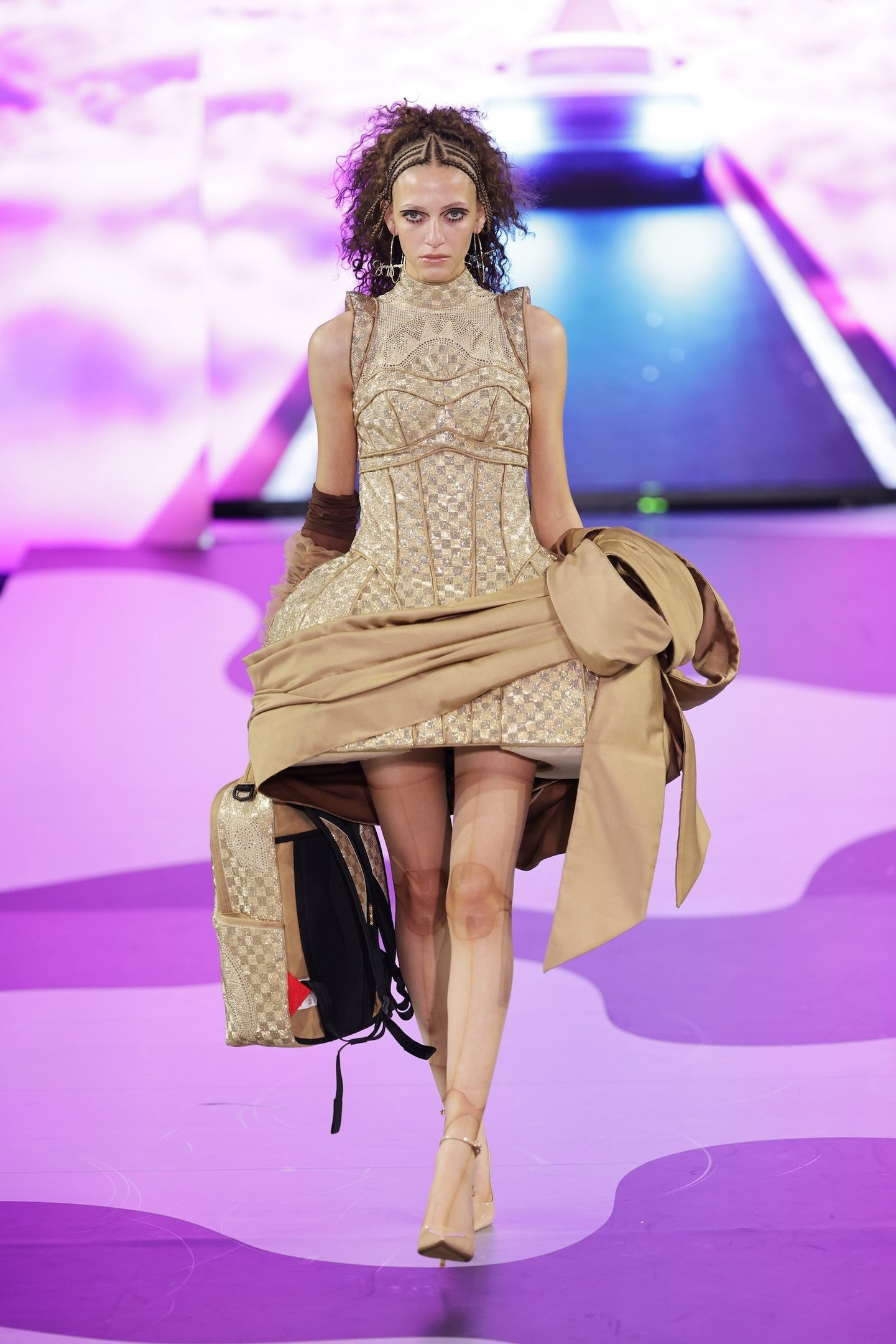 Modelo em vestido dourado com jaqueta e mochila combinando, desfilando em passarela na London Fashion Week SS26.