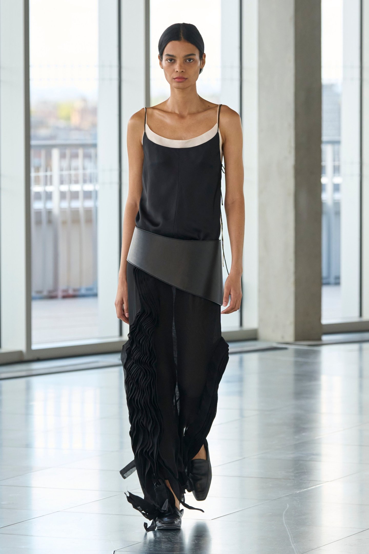 Modelo desfilando com roupa preta elegante durante a London Fashion Week SS26 em ambiente moderno e envidraçado.