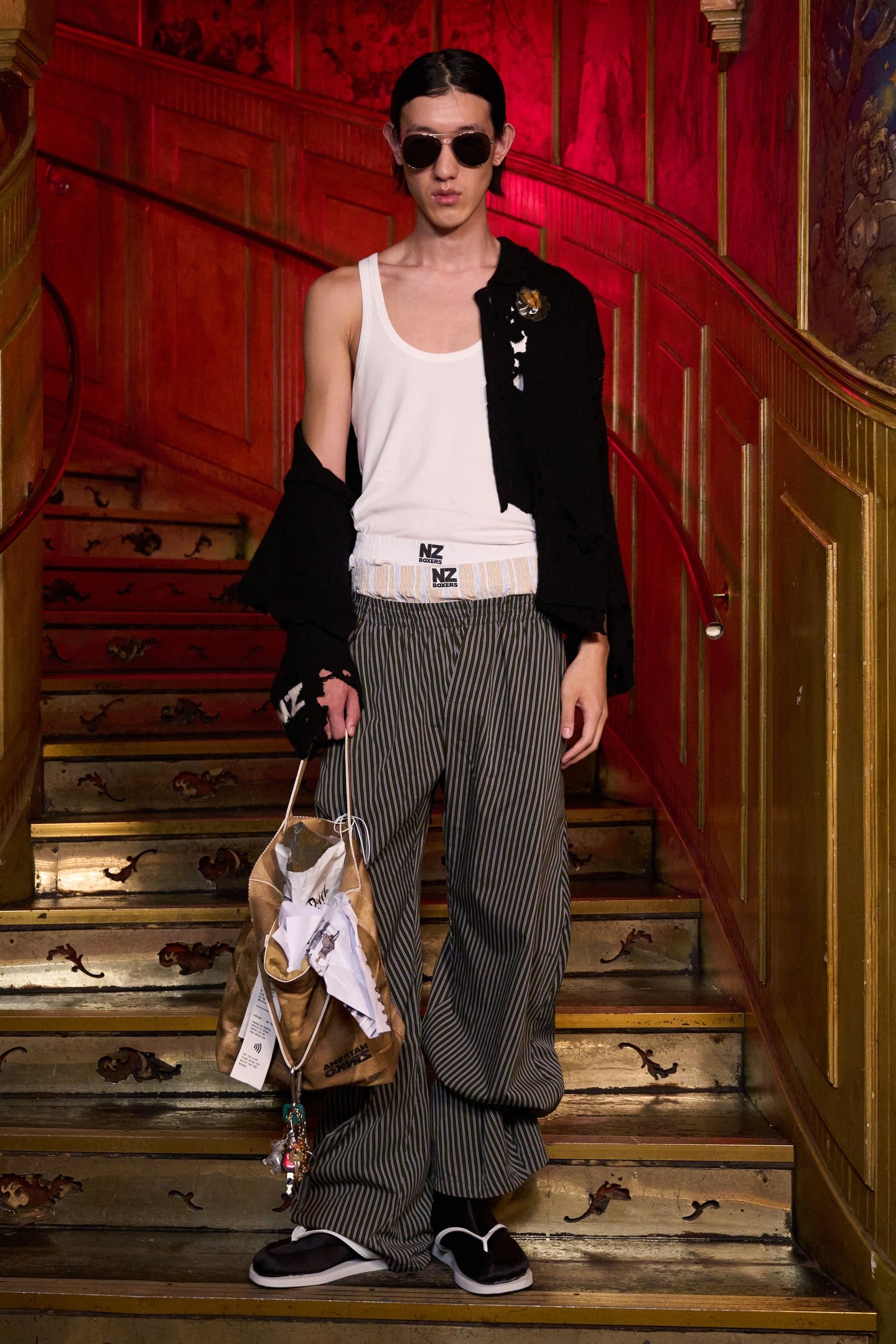 Modelo com top branco, calça listrada e óculos escuros no London Fashion Week SS26. Carrega bolsa marrom.