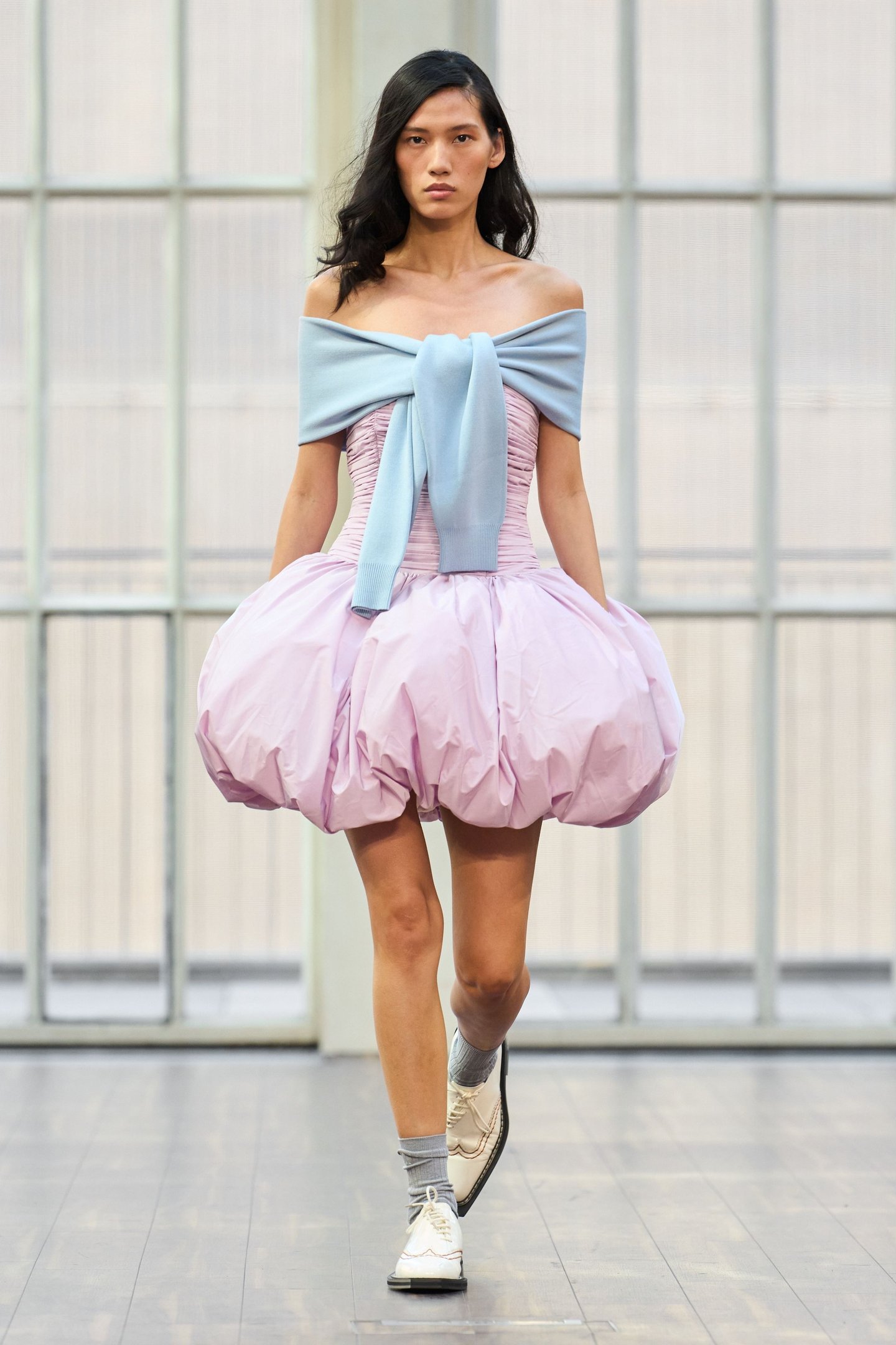 Modelo usando vestido rosa volumoso com suéter azul claro nos ombros, na London Fashion Week SS26.