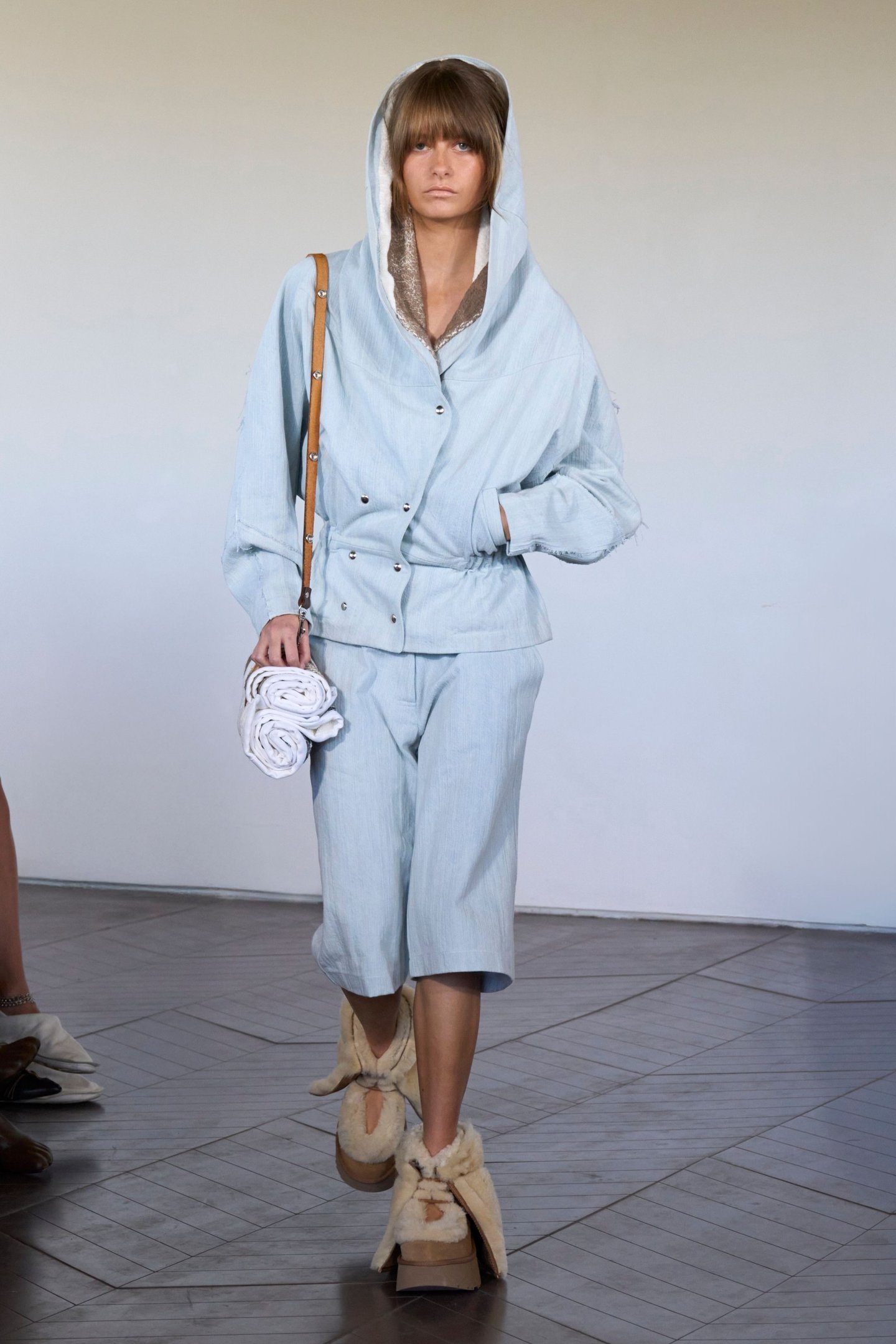 Desfile na London Fashion Week SS26, modelo usa conjunto azul claro com capuz e sandálias de pele.