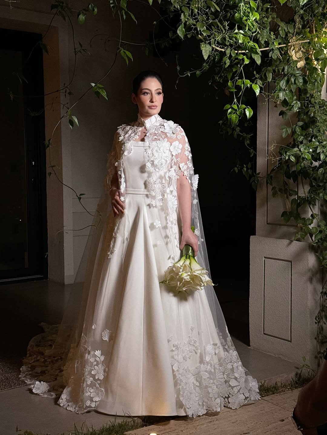 Noiva em vestido sofisticado com capa rendada, segurando buquê de flores brancas, ao lado de folhagens. Vestido Lucas Anderi.