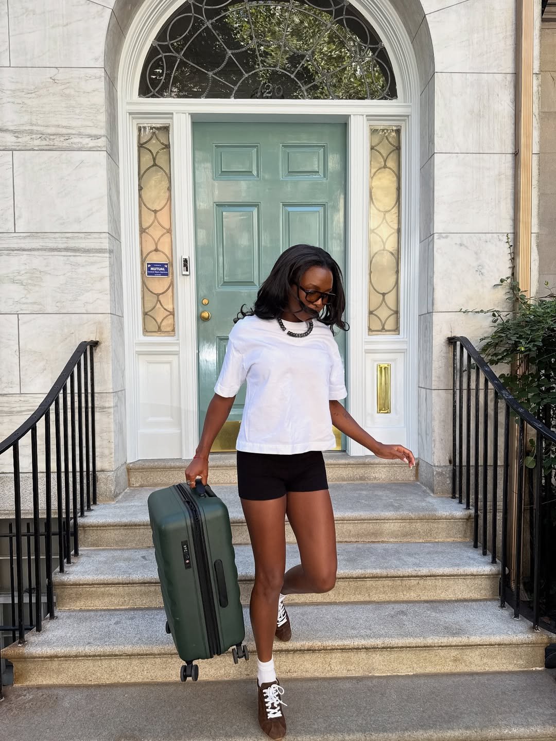 Mulher com mala verde em escada, usando camiseta branca, shorts pretos e tênis marrom feminino.