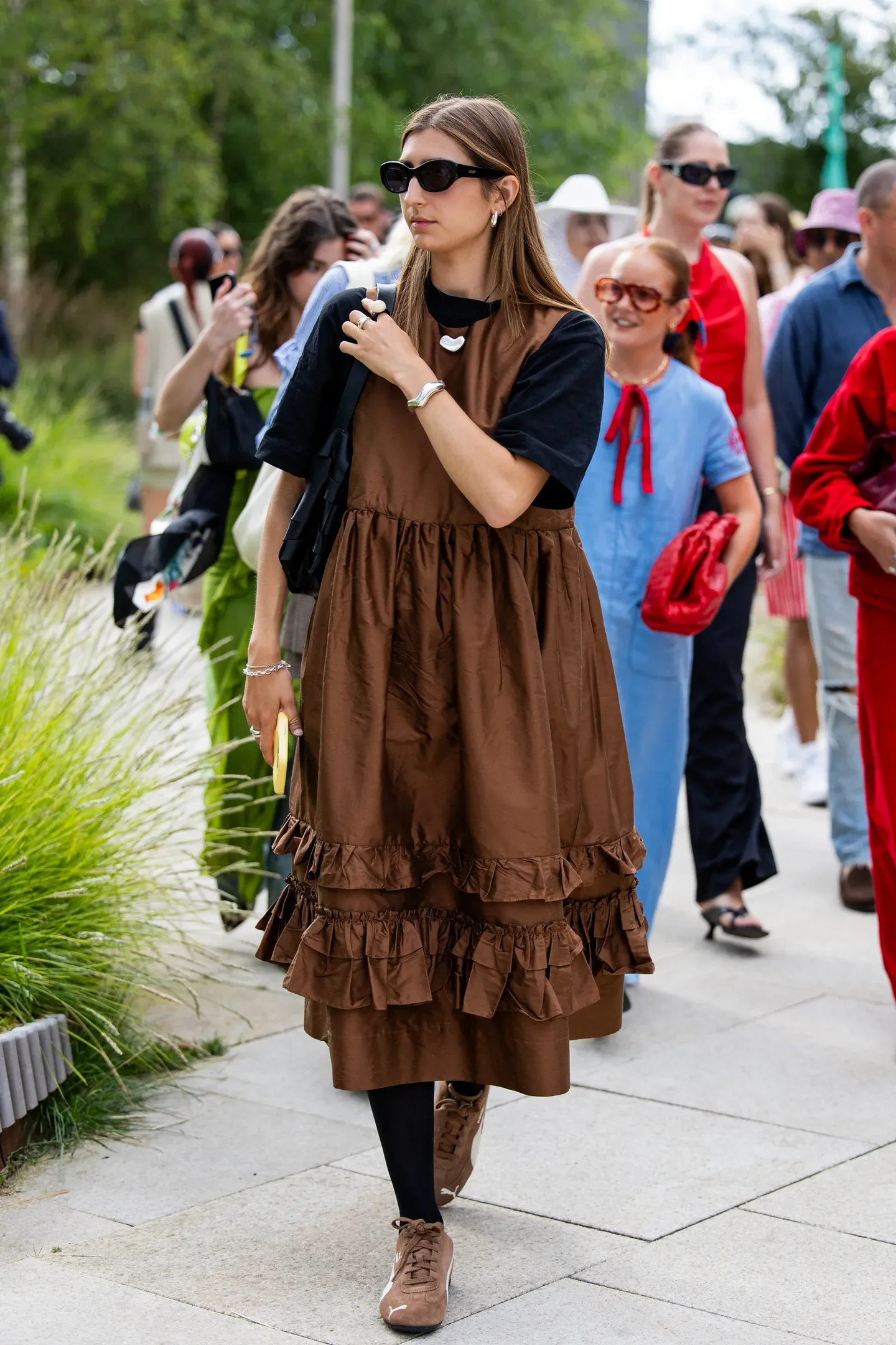 Mulher em vestido marrom com tênis marrom feminino, cercada por outras pessoas em um ambiente urbano.