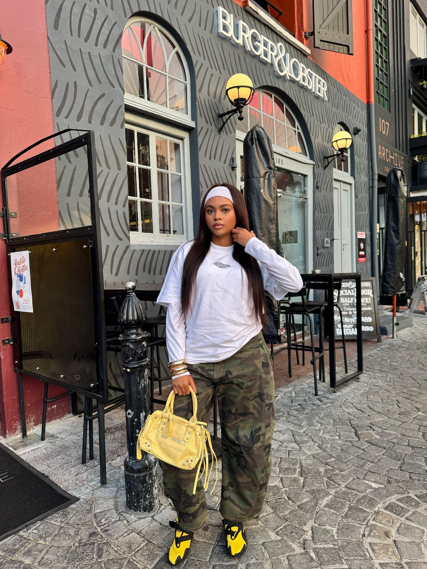 Pessoa com calça camuflada e blusa branca, segurando uma bolsa amarela. Tênis PUMA de tendência nos pés. Fundo de fachada de restaurante.