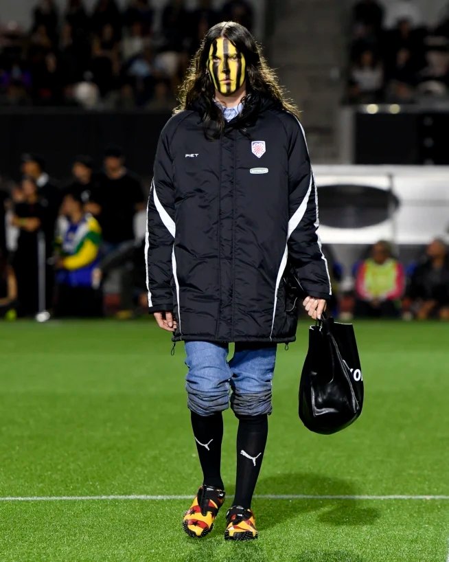 Pessoa usando casaco preto longo, calça jeans e tênis PUMA coloridos em uma passarela ao ar livre.