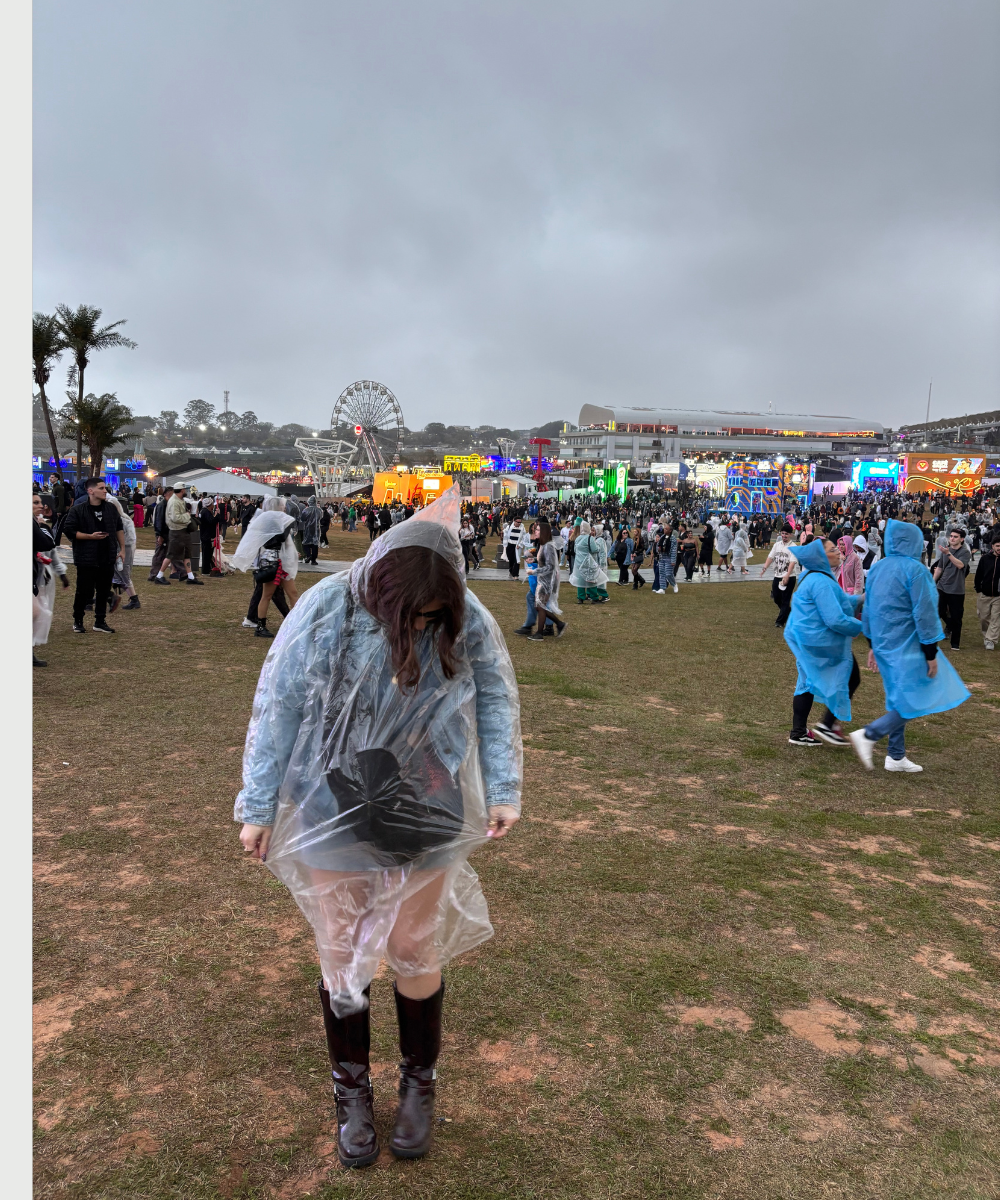Pessoas em um festival ao ar livre, com capas de chuva, próximo à roda-gigante. Evento The Town ao fundo.