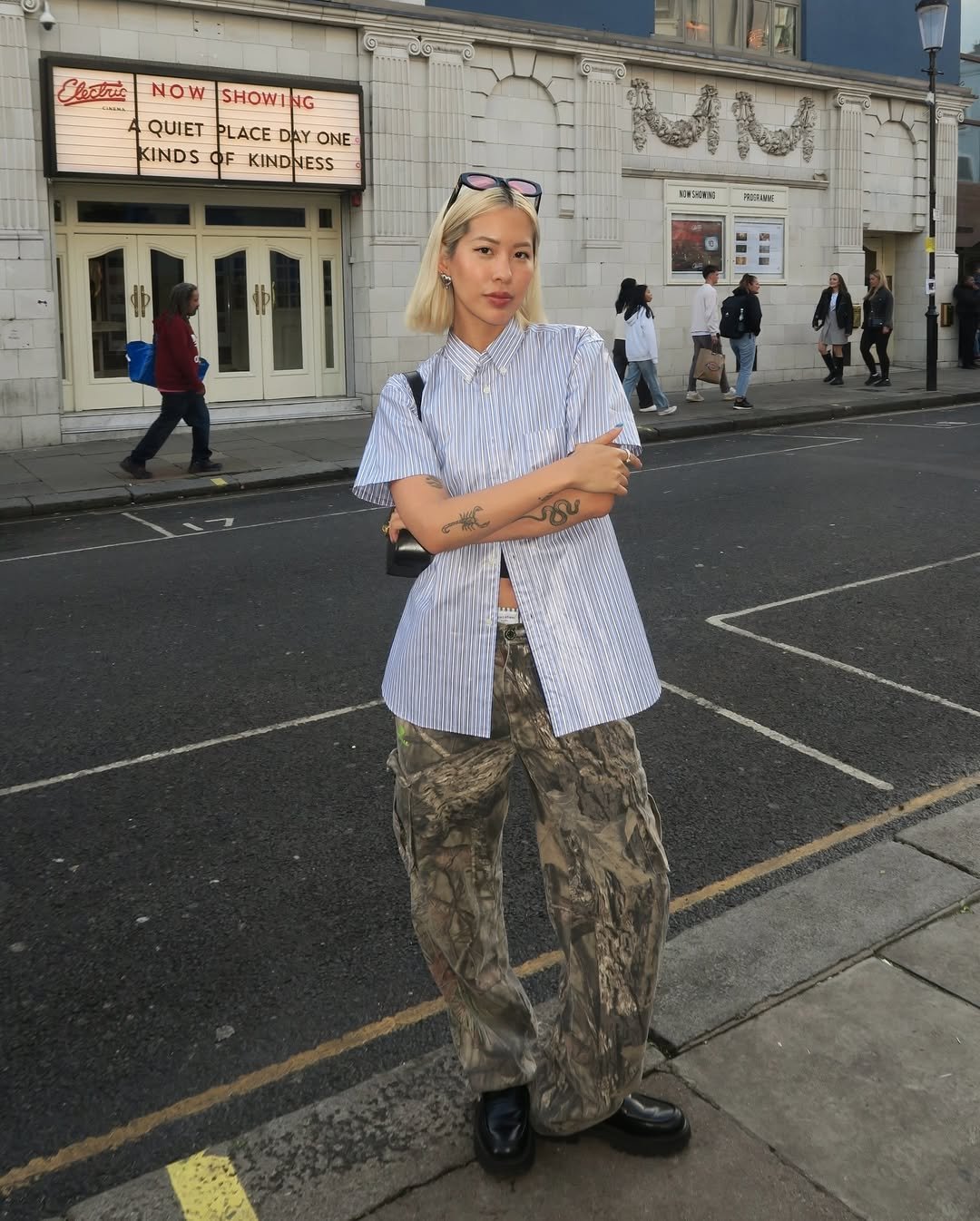 Pessoa em frente a um cinema, usando camisa listrada e calça camuflada, com pessoas ao fundo na calçada.